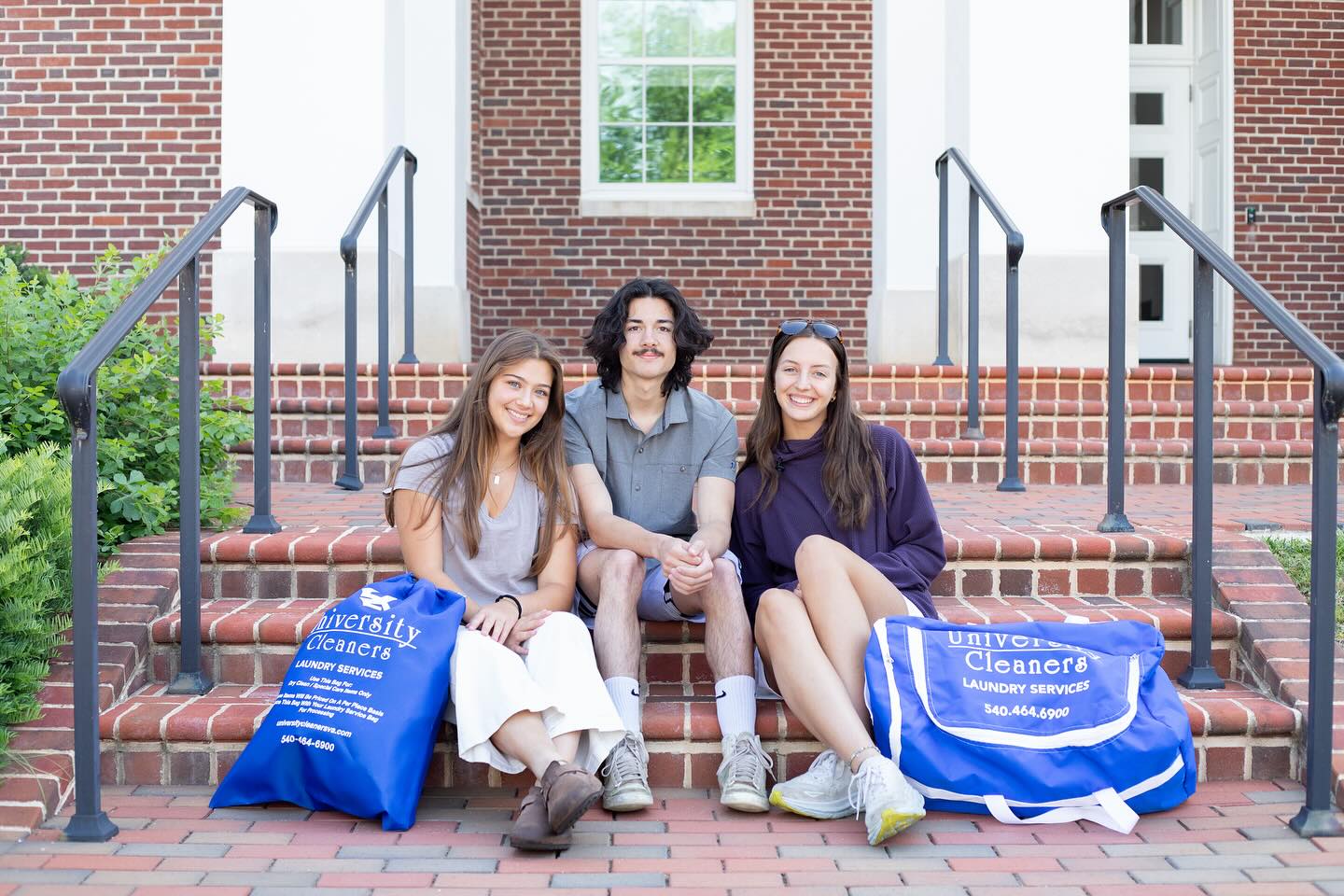 Three people sit on brick steps outside a building, smiling at the camera, with two blue University Cleaners laundry bags beside them.
