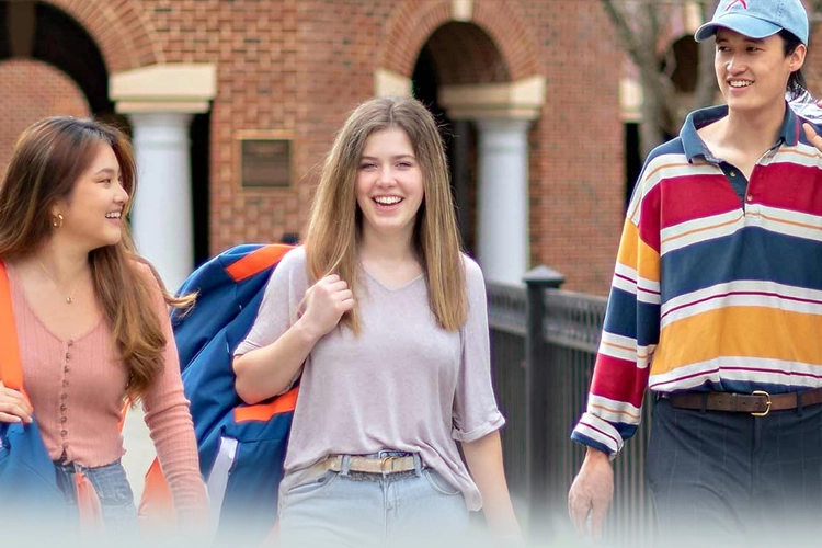 Three young adults walk together outside a brick building, smiling and talking. One carries a jacket over their shoulder.