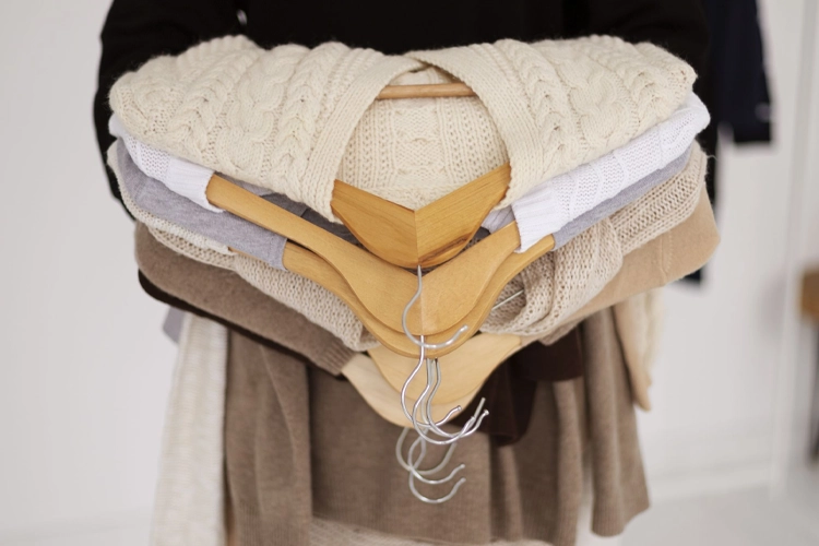 Person holding a stack of folded knit sweaters in neutral colors with wooden hangers resting on top.