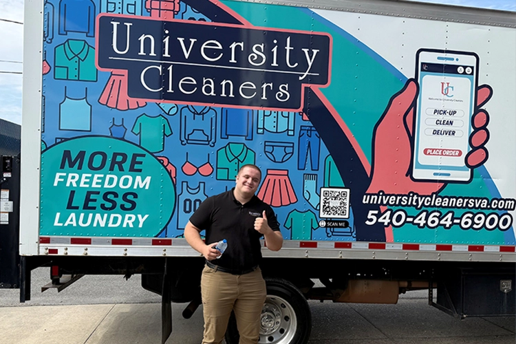 A person stands smiling and giving a thumbs-up in front of a University Cleaners delivery truck with service details and a colorful laundry-themed design.