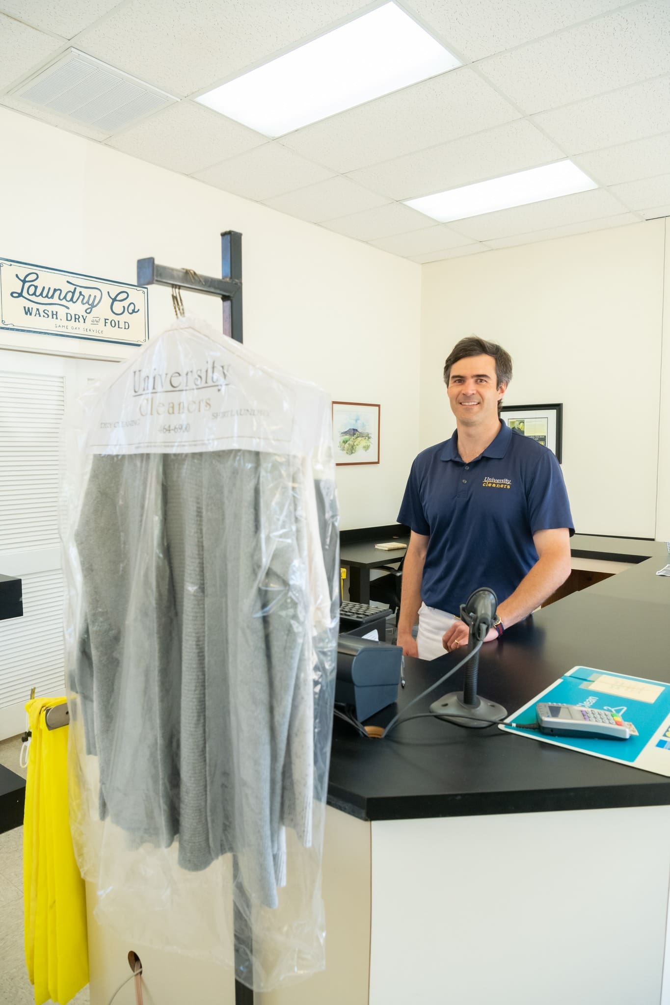 A man stands behind a counter at a dry cleaning shop with a garment in plastic hanging in the foreground and a computer, folder, and pen on the counter.