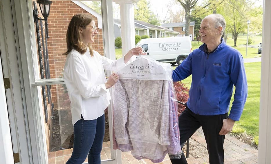 A woman receives freshly cleaned clothes in a plastic cover from a deliveryperson at her doorstep, with a University Cleaners van parked outside.