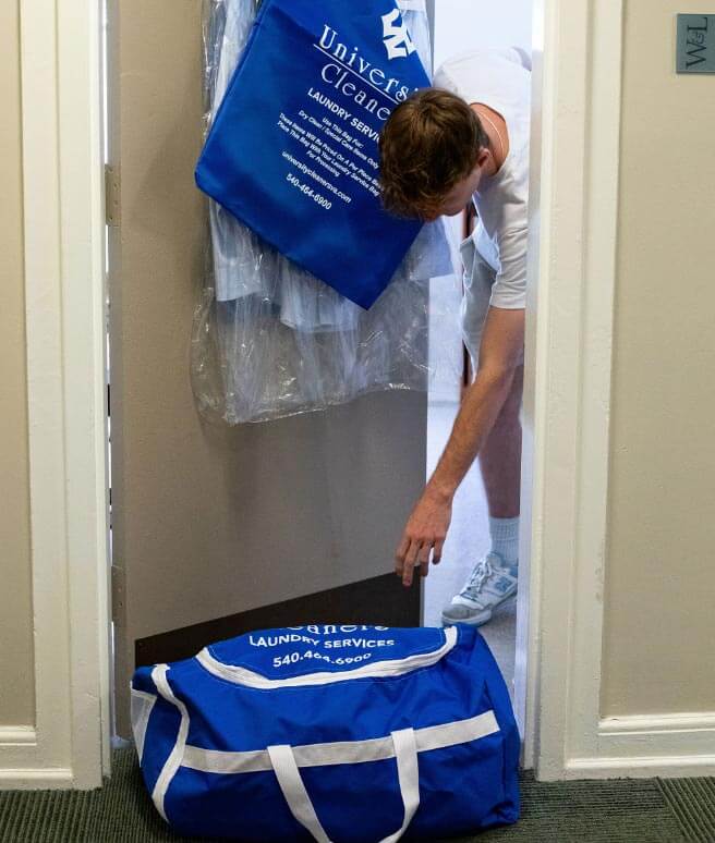 A person collects laundry from a doorstep, with a blue laundry service bag and a hanging garment bag visible.