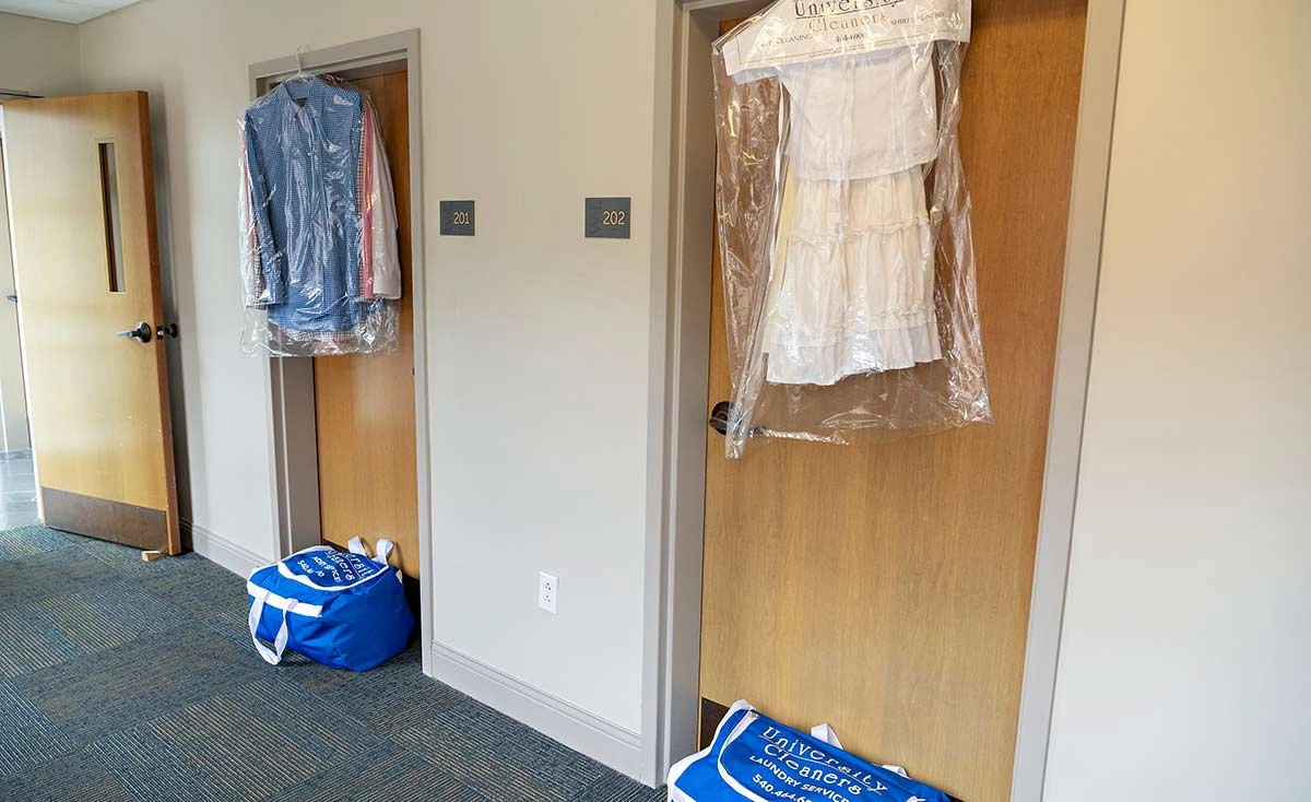 Two dorm room doors with dry cleaning bags hanging on them and laundry bags placed on the floor outside each door in a hallway.