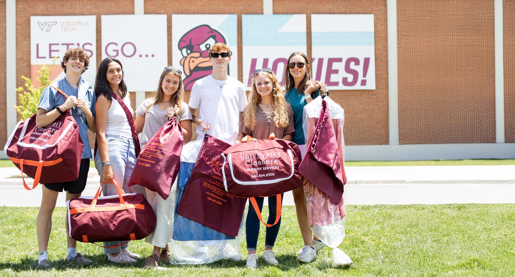 Six young adults stand outside Virginia Tech holding maroon laundry bags, with a building and Hokies signage in the background.