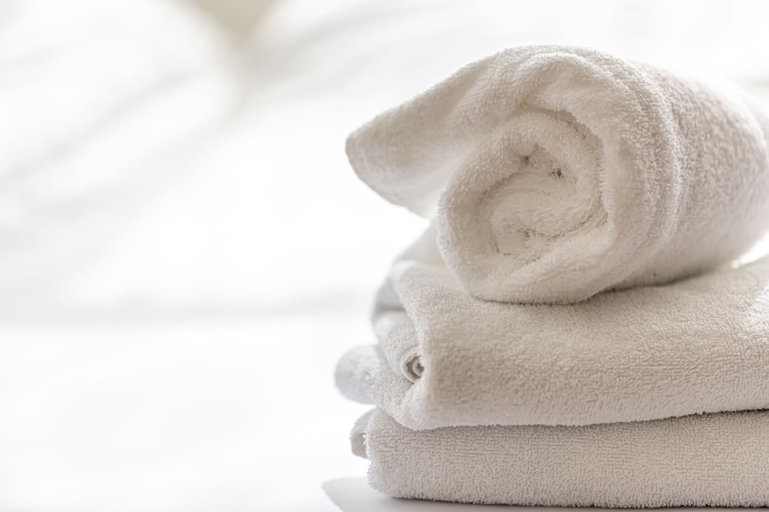 Three neatly folded white towels stacked on a white surface with a soft, blurred background.