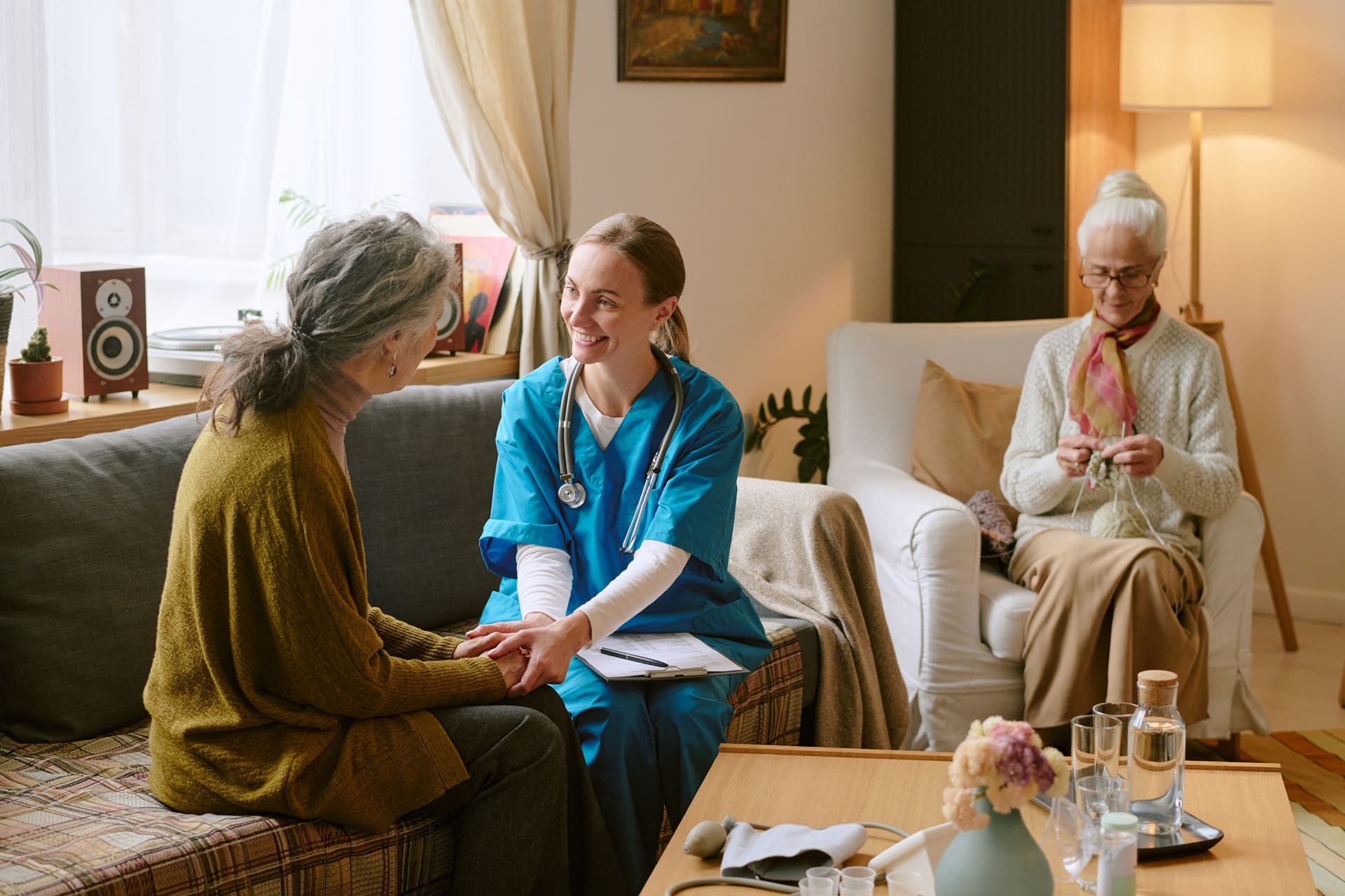 A nurse in blue scrubs speaks with an older woman on a couch while another elderly woman knits in the background in a cozy living room.