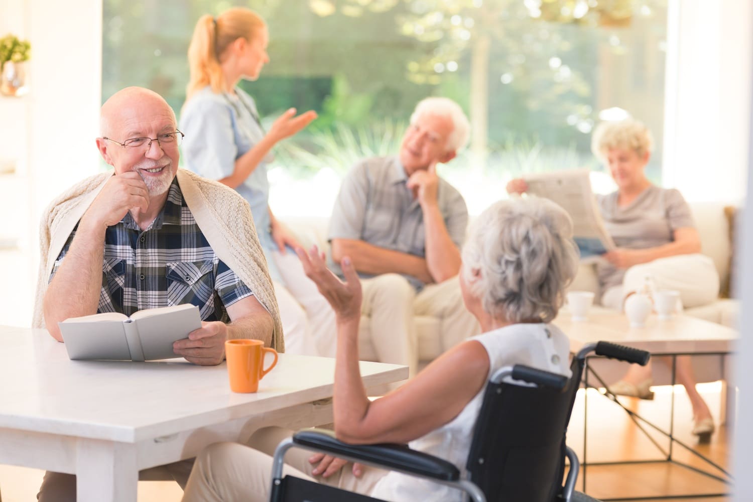 A man sits at a table with a book and coffee, talking to a woman in a wheelchair. Three older adults and a caregiver converse in the background in a bright room.