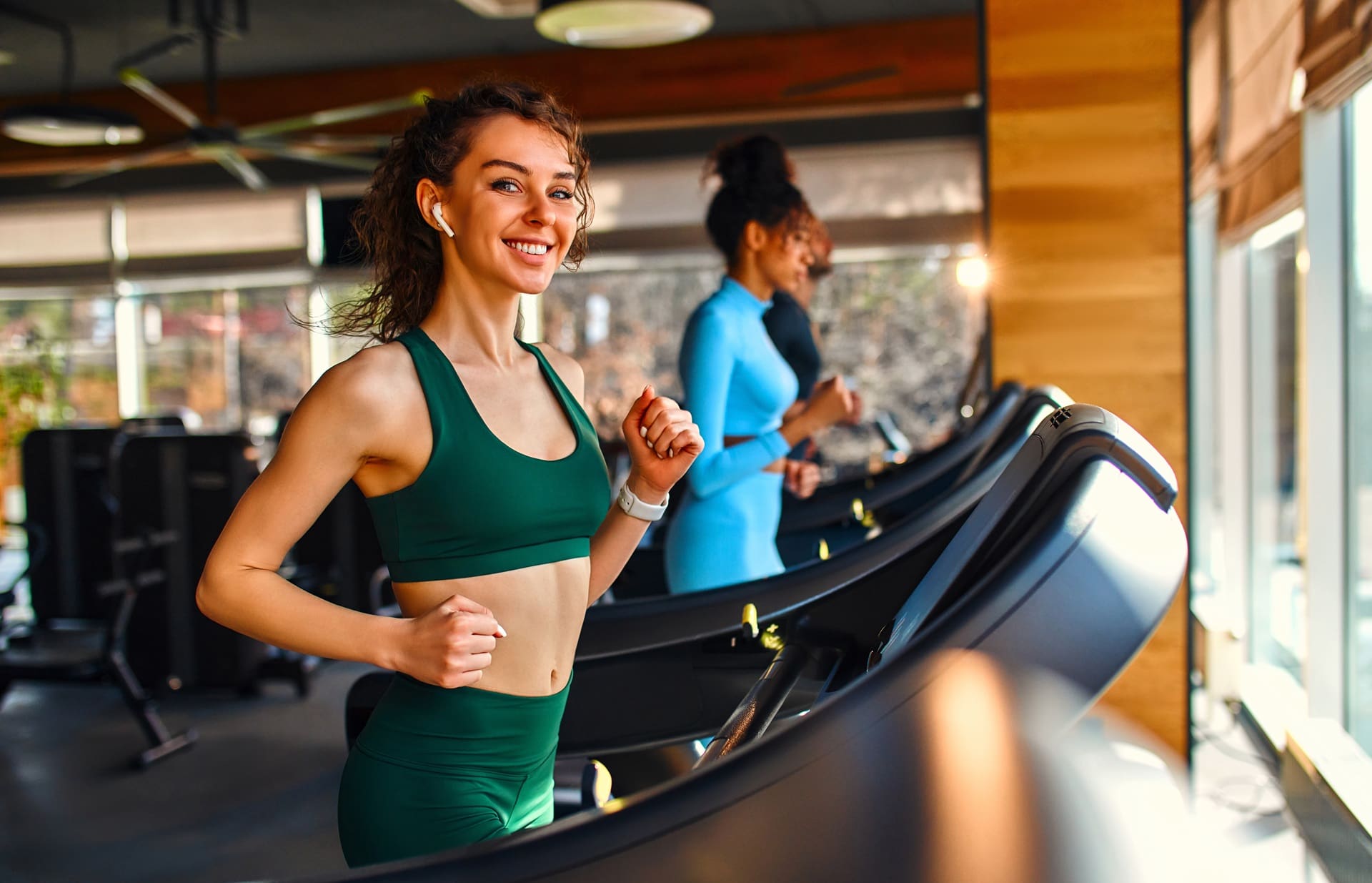Three women exercise on treadmills in a modern gym, with one woman in green athletic wear smiling at the camera.