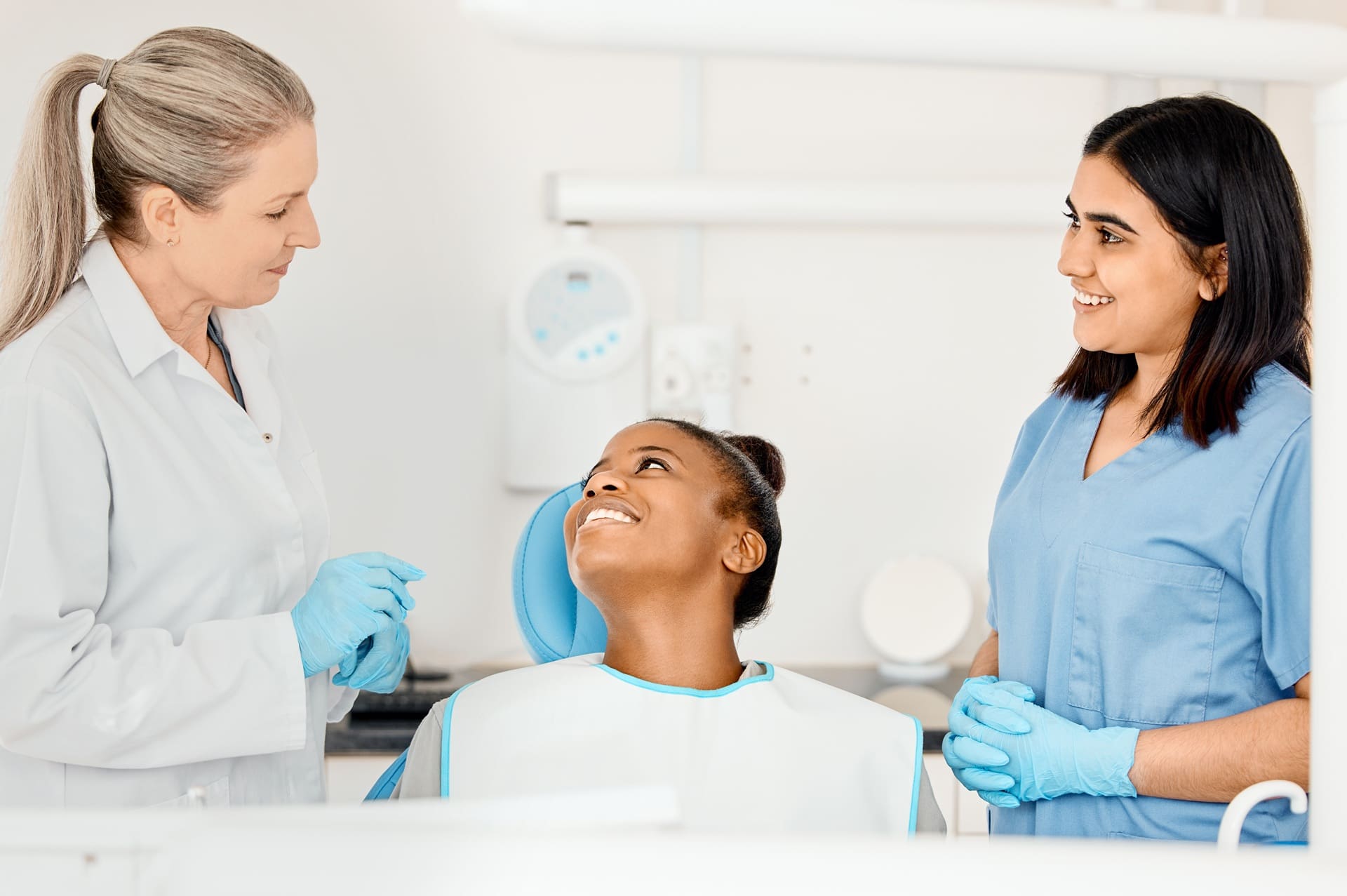 A dental patient sits in a chair smiling at a dentist and a dental assistant, both wearing gloves and uniforms in a clinical setting.