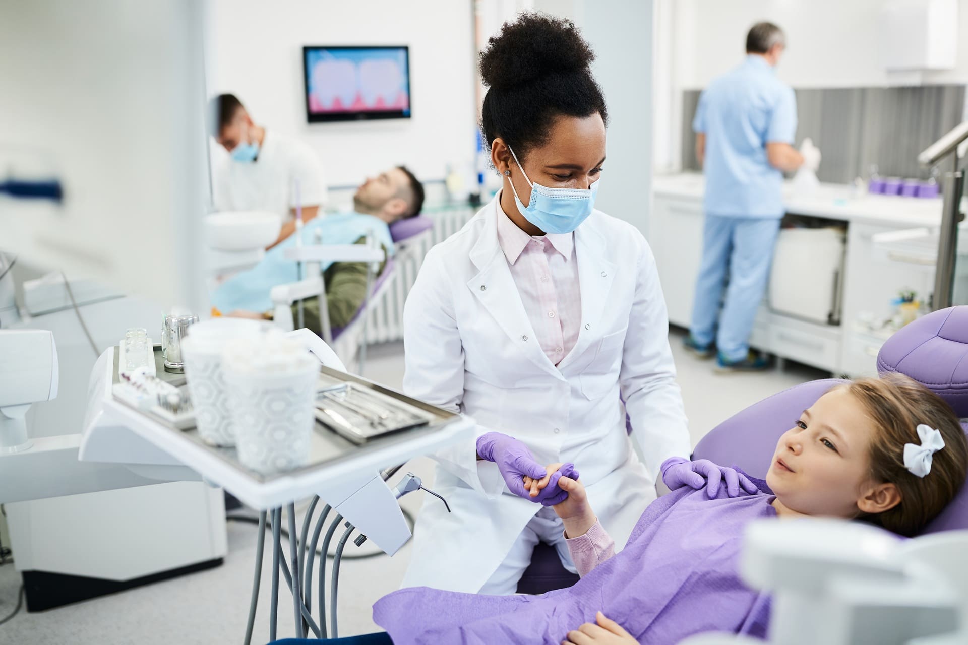 A dentist wearing a mask and gloves talks to a young girl in a dental chair, while other patients and staff are visible in the background of the clinic.
