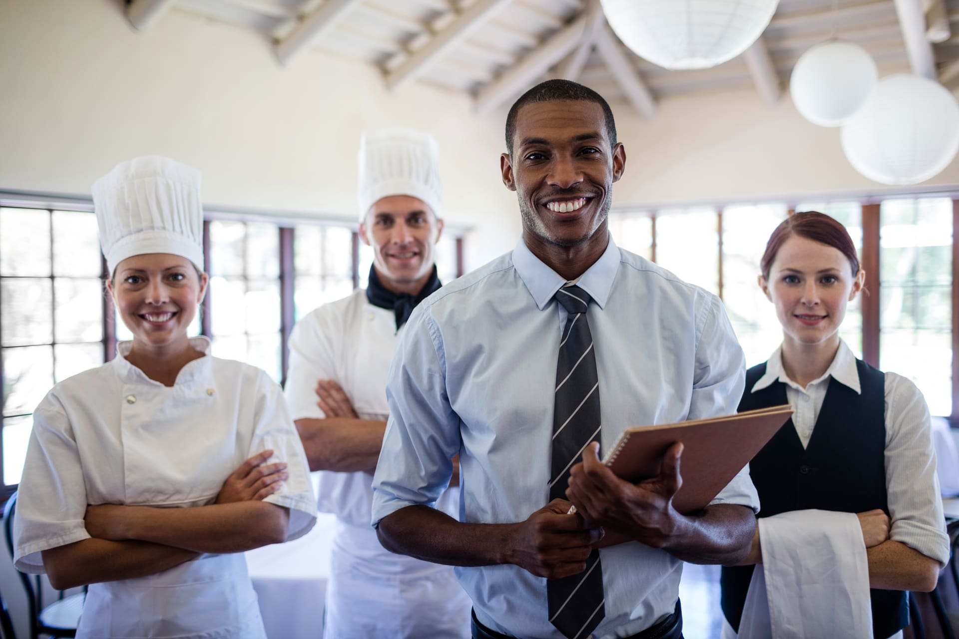A restaurant manager holding a clipboard stands in front of three staff members, including two chefs in uniform and a server with a towel.