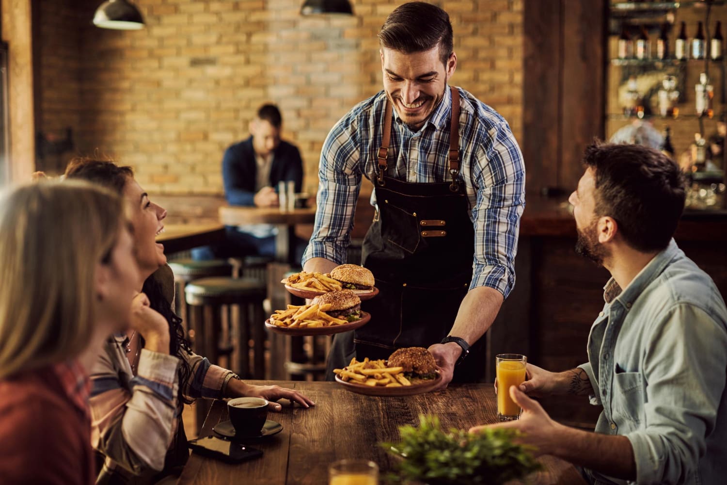 A waiter in a plaid shirt and apron serves plates of burgers and fries to three people seated at a wooden restaurant table.