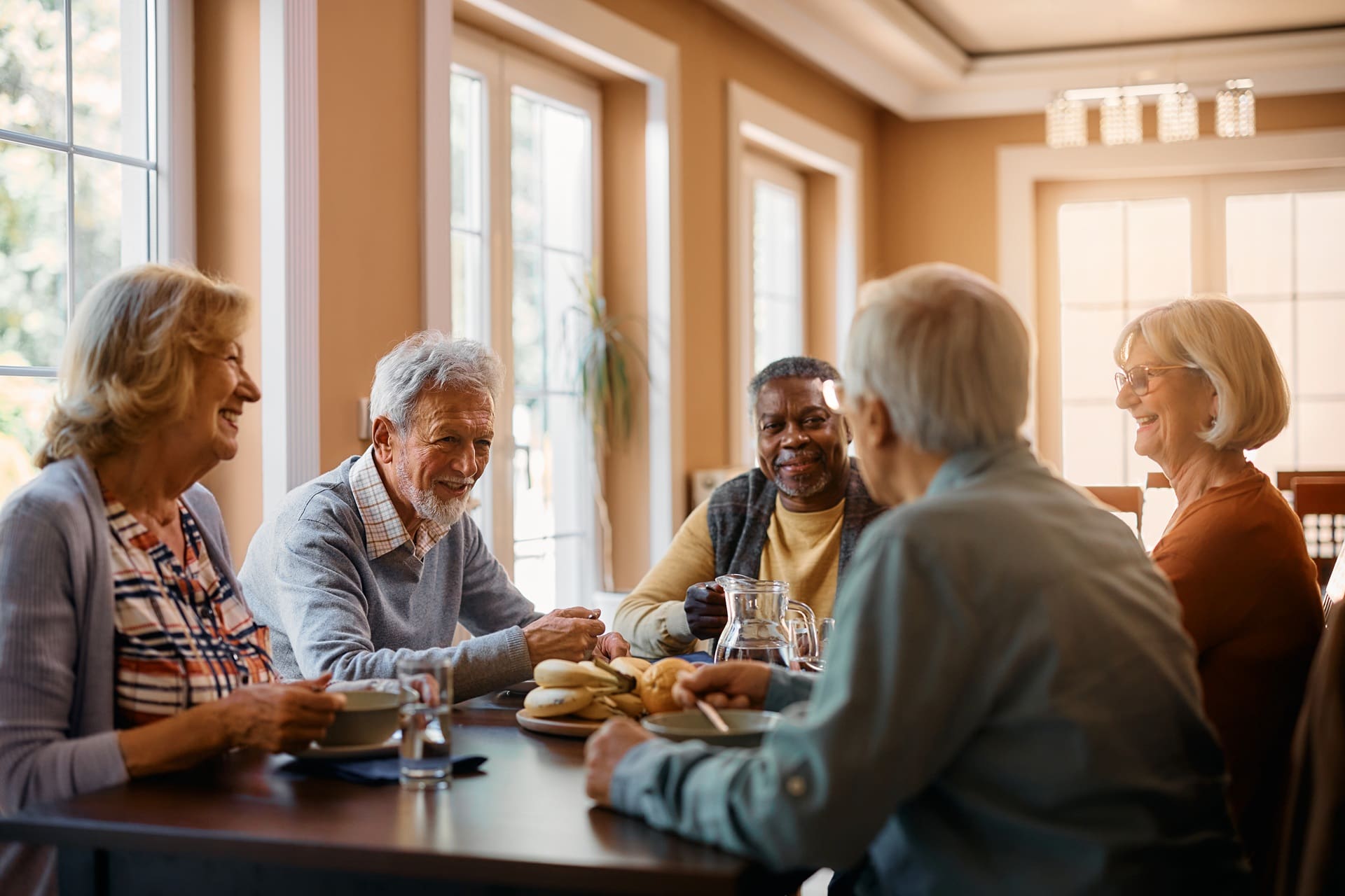 Five older adults are sitting around a dining table, smiling and conversing, with plates, cups, and a jug of water in a well-lit room.