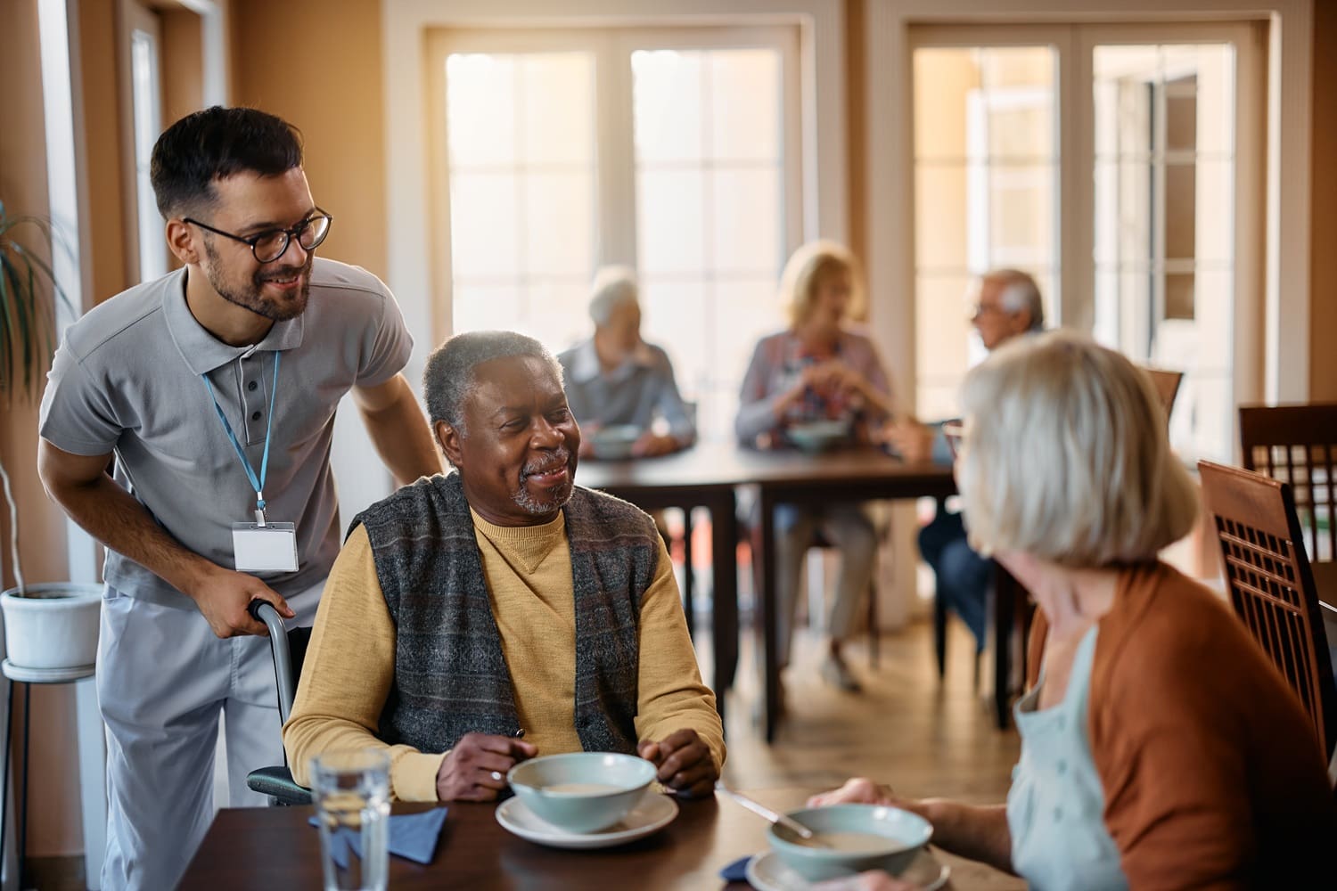 A caregiver stands beside an older man in a wheelchair at a dining table, talking to a woman. Other seniors are seated and conversing in the background.