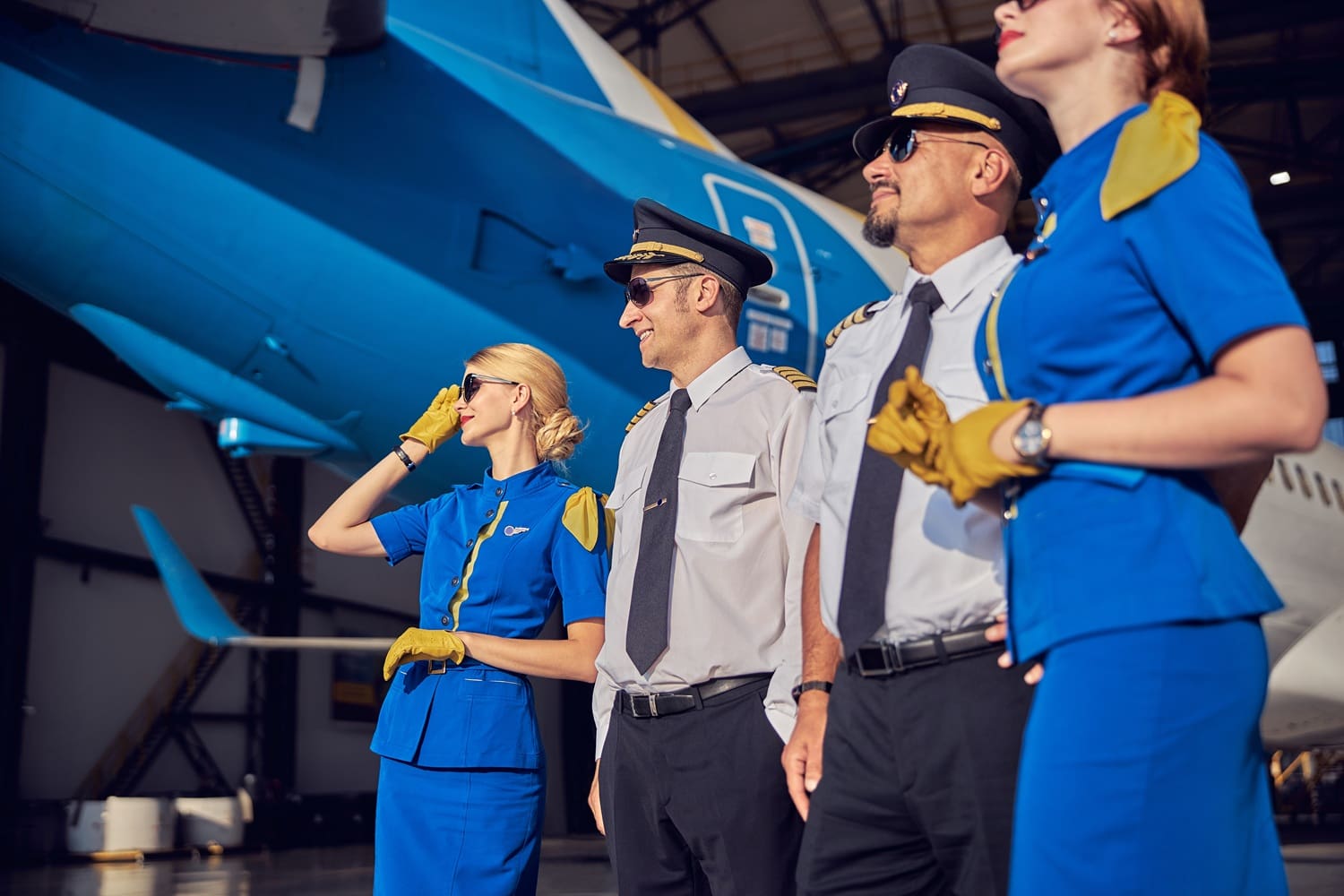 Four airline crew members, two in blue uniforms and two in pilot uniforms, stand together in front of a large airplane inside a hangar.