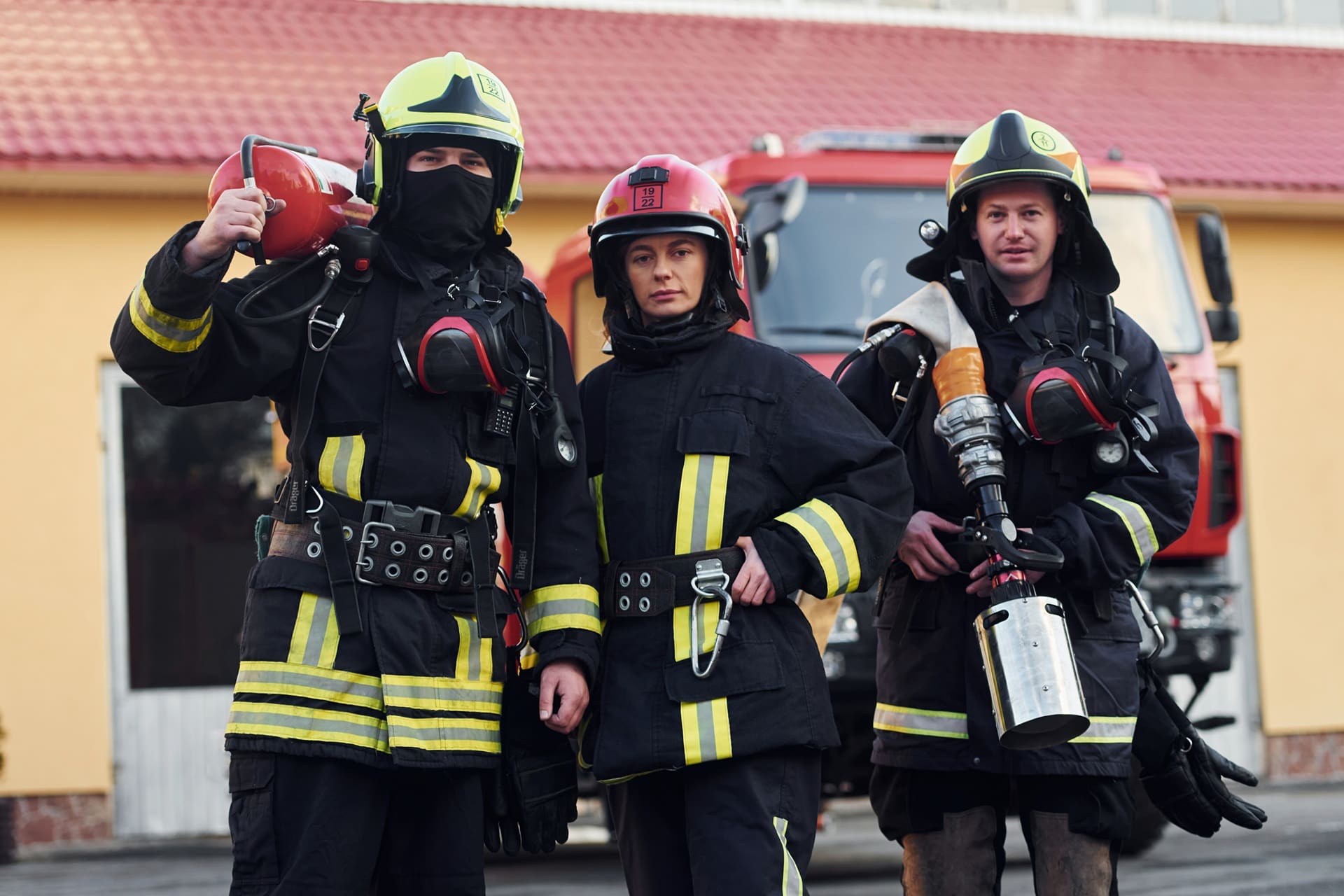 Three firefighters in full gear stand in front of a fire truck, holding equipment and looking at the camera.