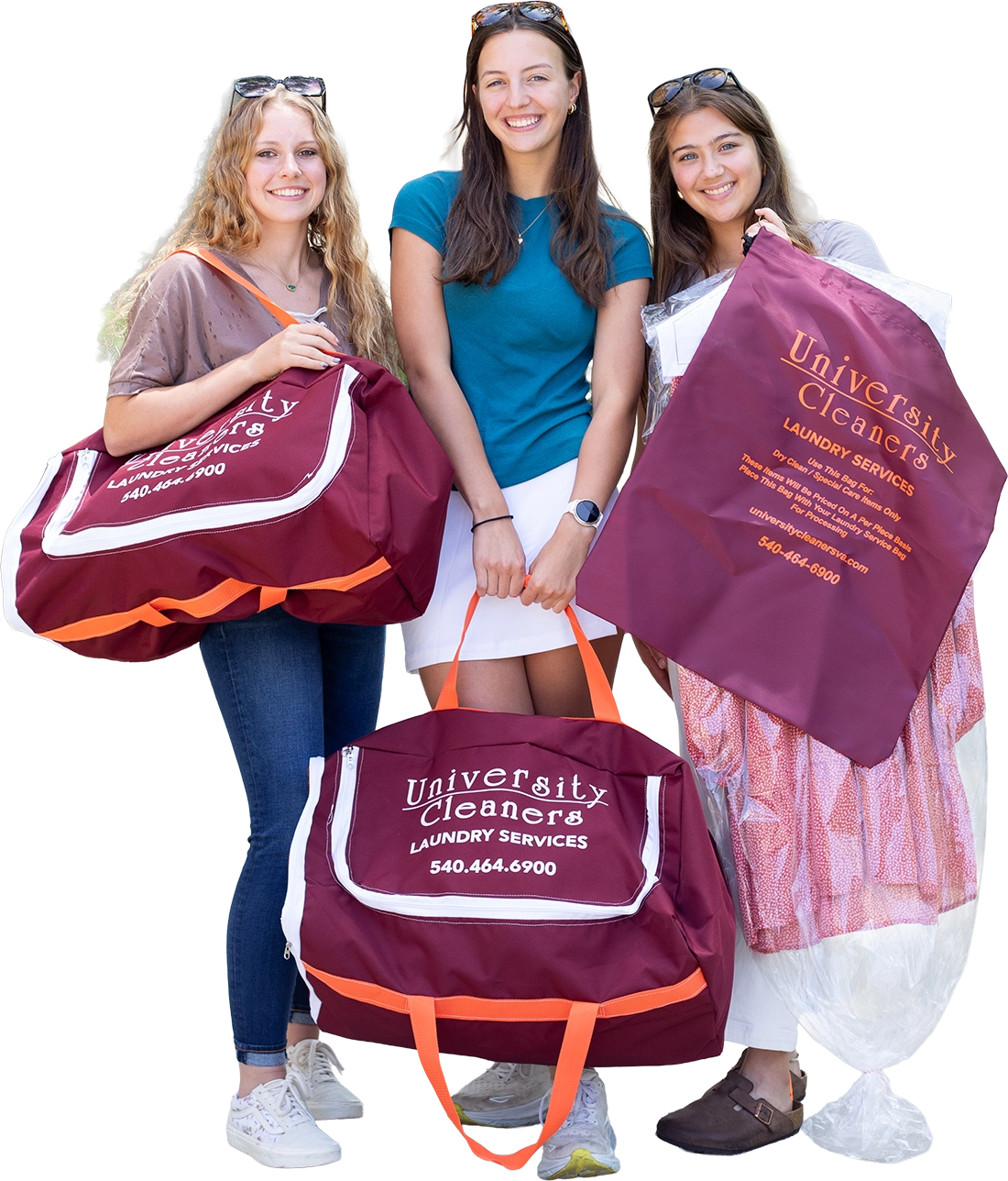 Three young women stand smiling, each holding maroon University Cleaners laundry bags with laundry service contact information printed on them.