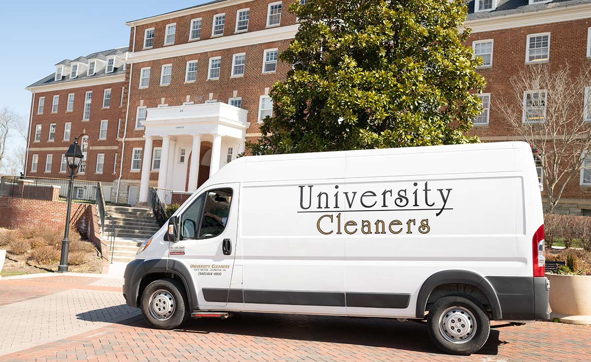 A white van labeled "University Cleaners" is parked in front of a brick university building with columns and several windows.