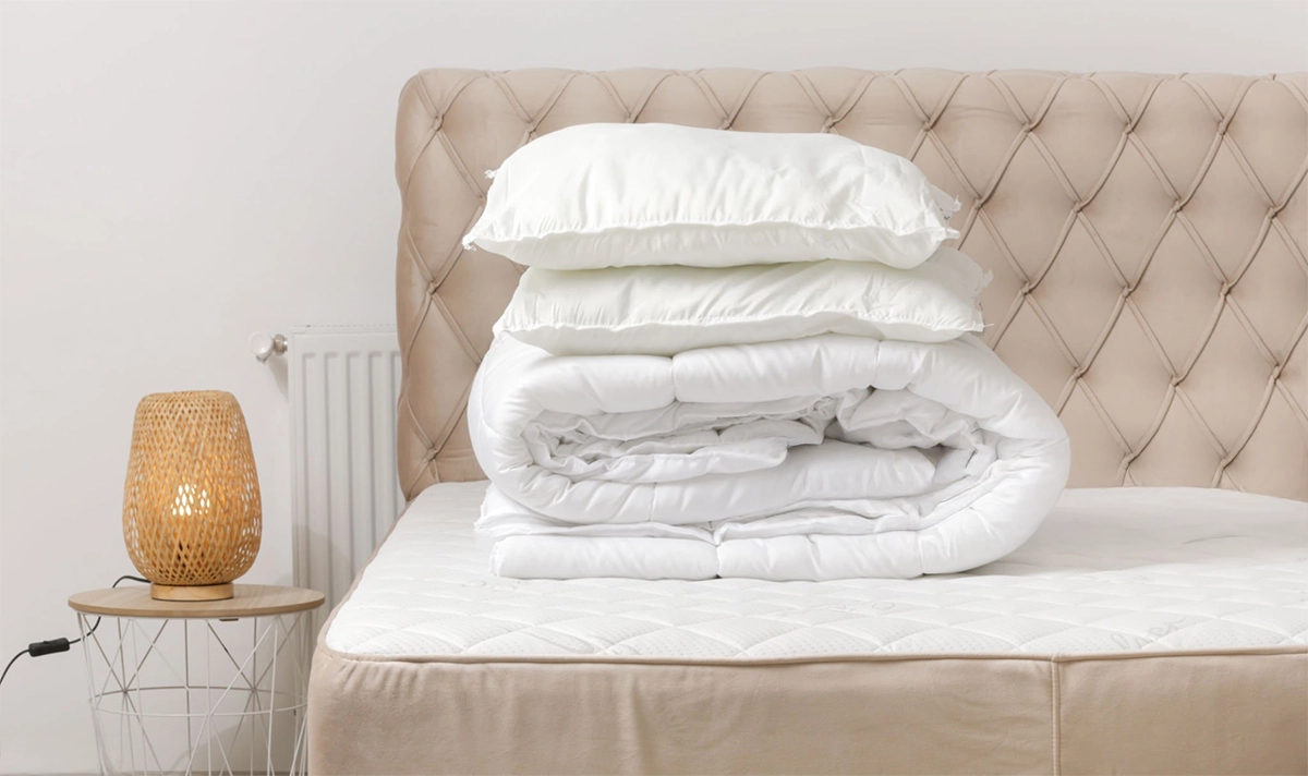 A bed with a tufted beige headboard, a mattress, neatly folded white bedding, and pillows beside a small table with a woven lamp.