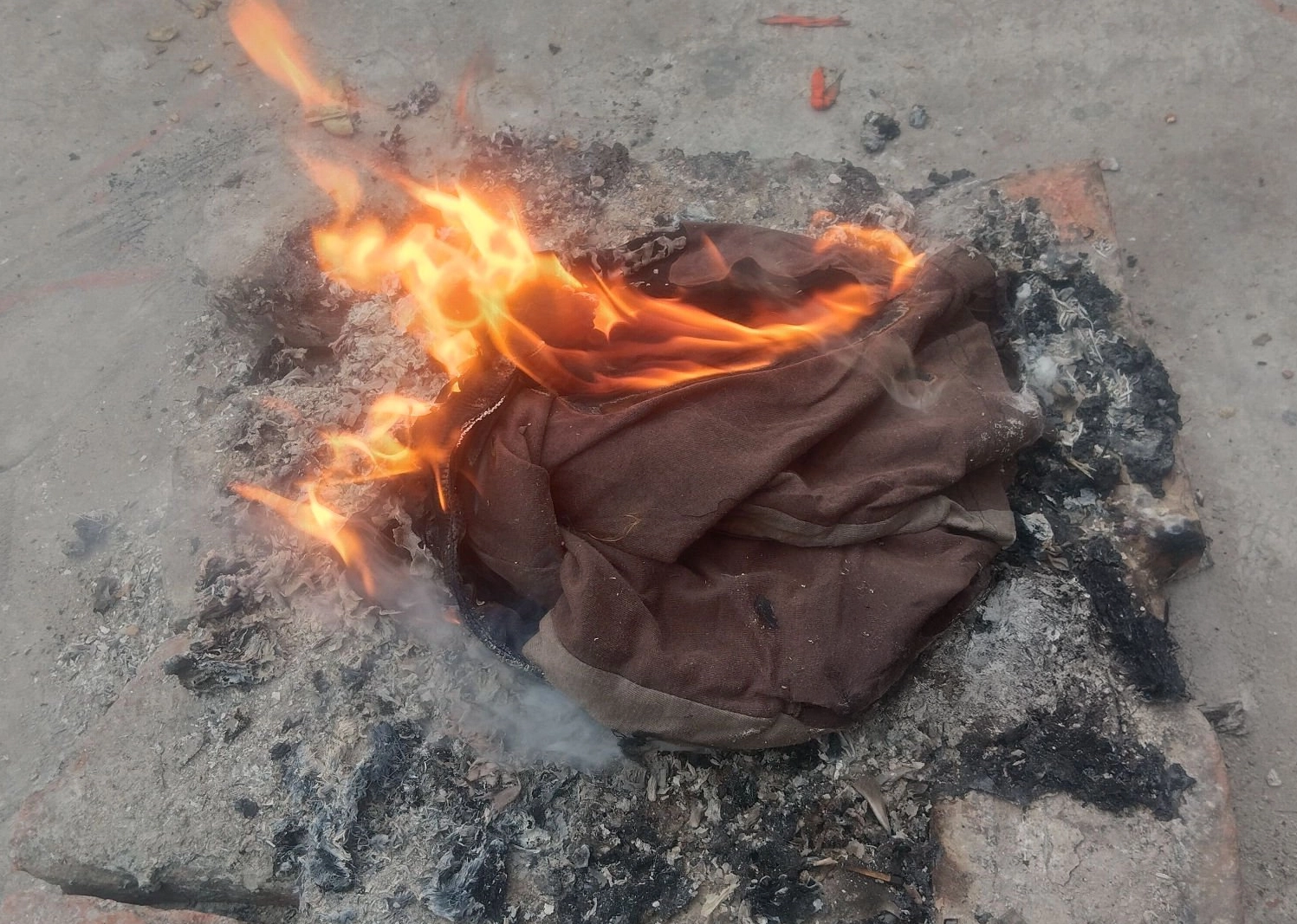 A piece of brown fabric is burning with visible flames and smoke, placed on a pile of ash and bricks on a concrete surface.