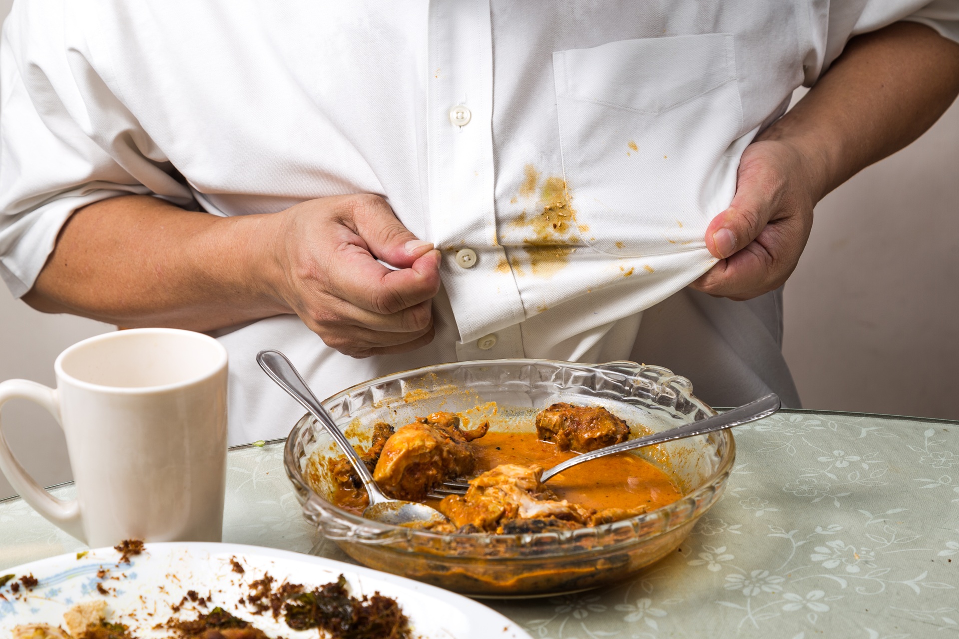 A person in a white shirt examines a food stain on their chest while sitting at a table with a dish of curry, a mug, and a plate with food remnants.