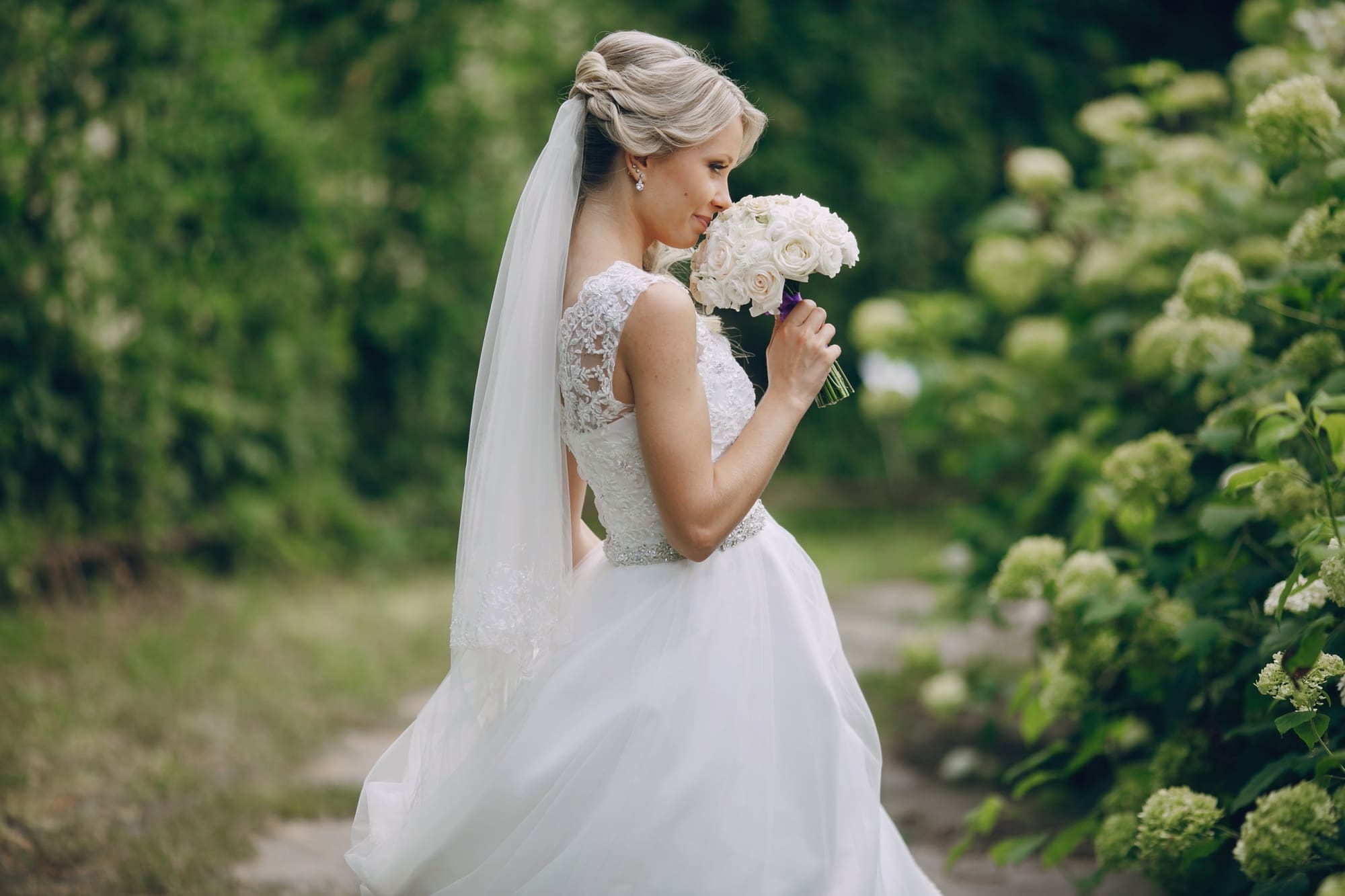 A bride in a white wedding dress and veil holds a bouquet of white roses, standing outdoors near green bushes.