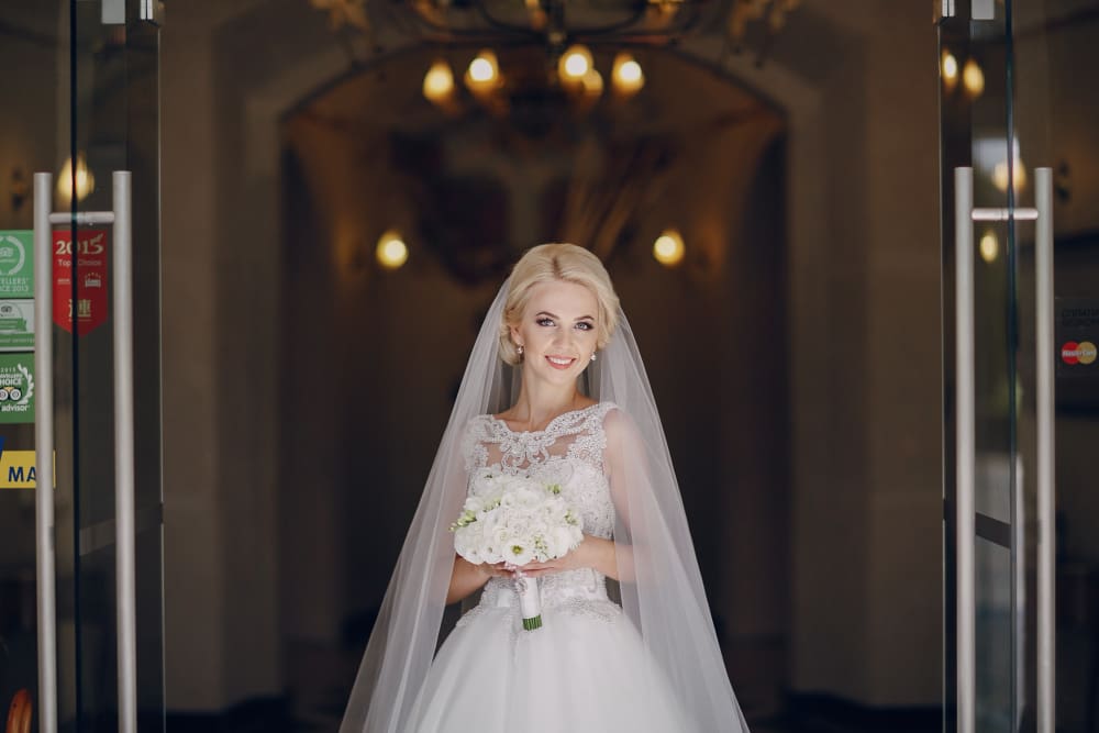 A bride in a white wedding dress and veil stands indoors, holding a bouquet of white flowers and smiling at the camera.