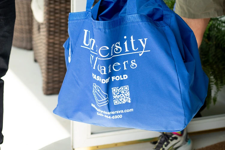 A person carries a blue University Cleaners laundry bag with white text, a QR code, and contact information printed on it.