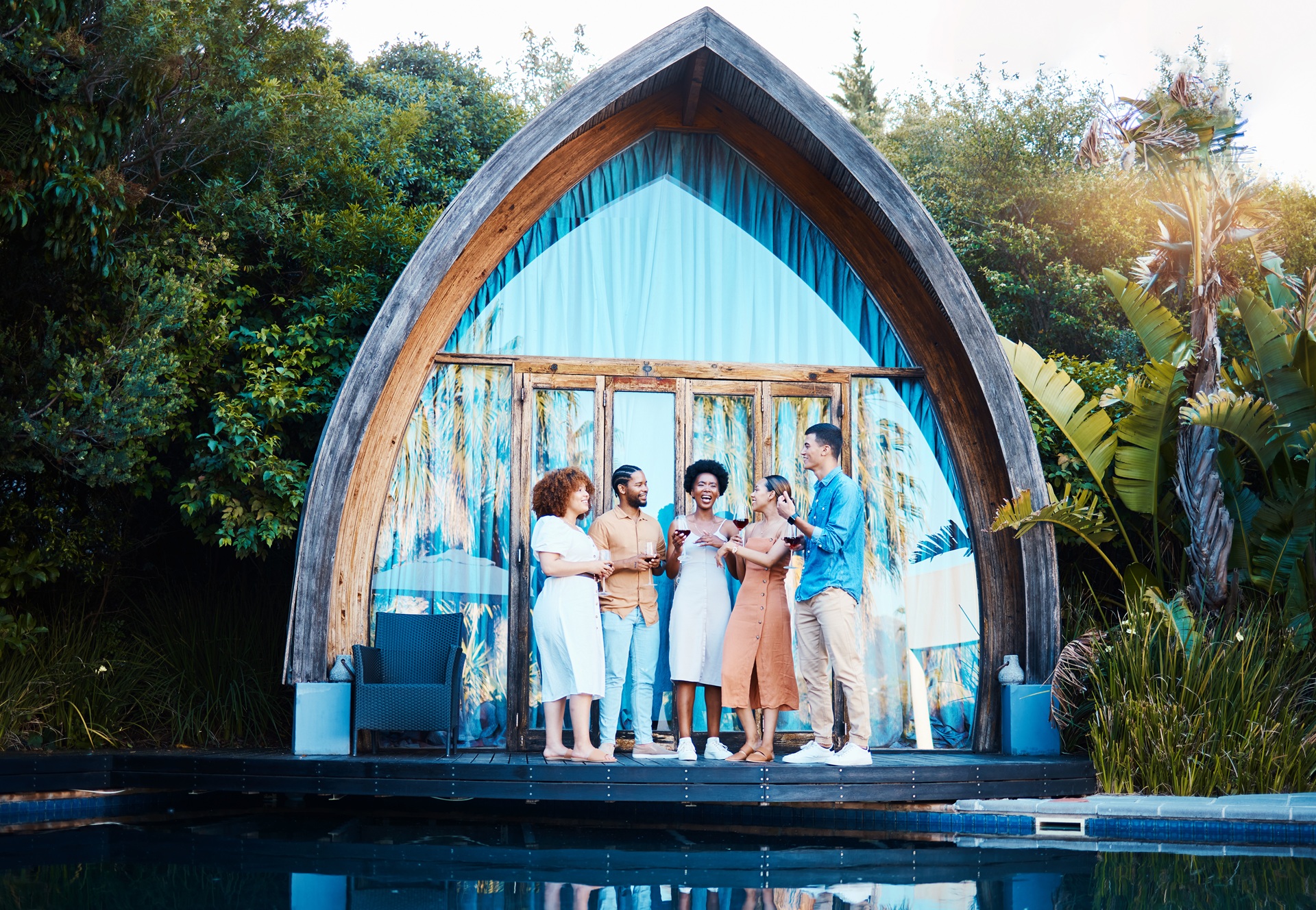Six people stand together on a wooden deck by a pool, talking and holding drinks in front of a small arched cabin surrounded by lush greenery.