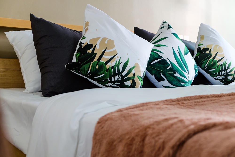 A neatly made bed with white sheets, black and tropical leaf print pillows, and a soft brown blanket.