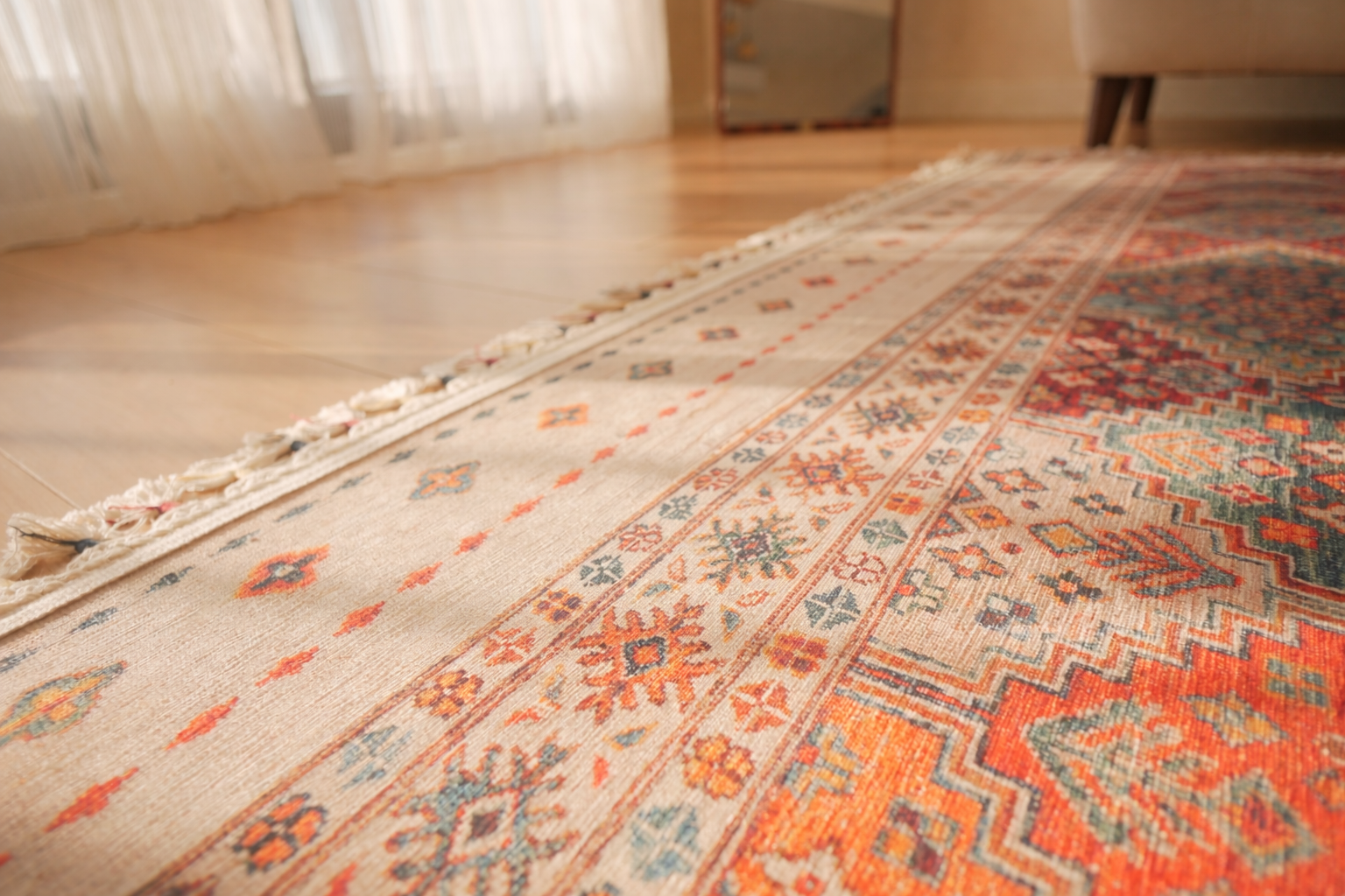 Close-up of a patterned rug with orange, blue, and beige designs on a light wooden floor near a window with sheer curtains.