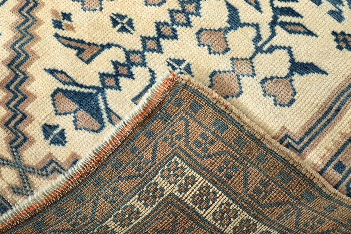 Close-up of a patterned rug with one corner folded over, showing both the front design in blue and beige and the back with muted colors and visible stitching.