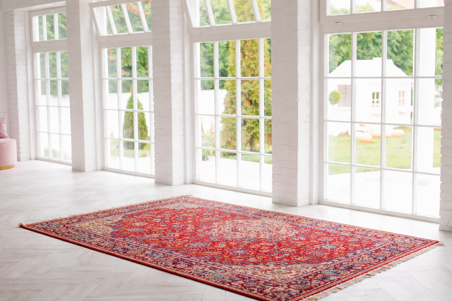 A large red patterned rug is laid on a white floor in a bright room with tall windows and views of a green garden outside.