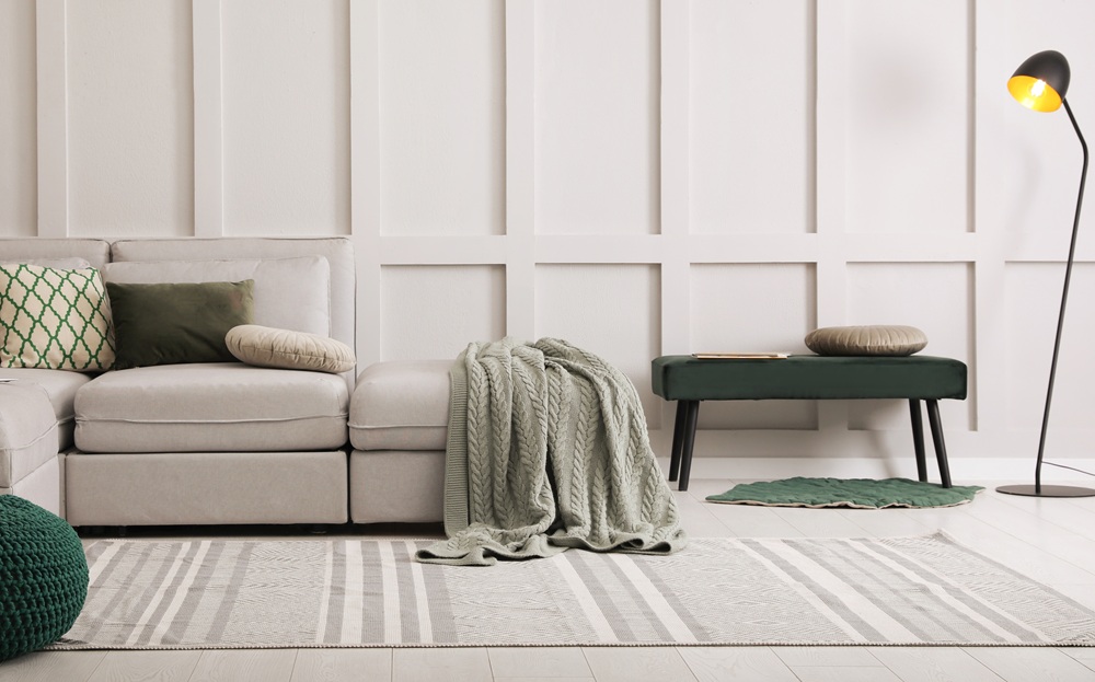 Modern living room with a light gray sectional sofa, green pillows, blanket, bench, floor lamp, and a striped rug on light wood flooring.