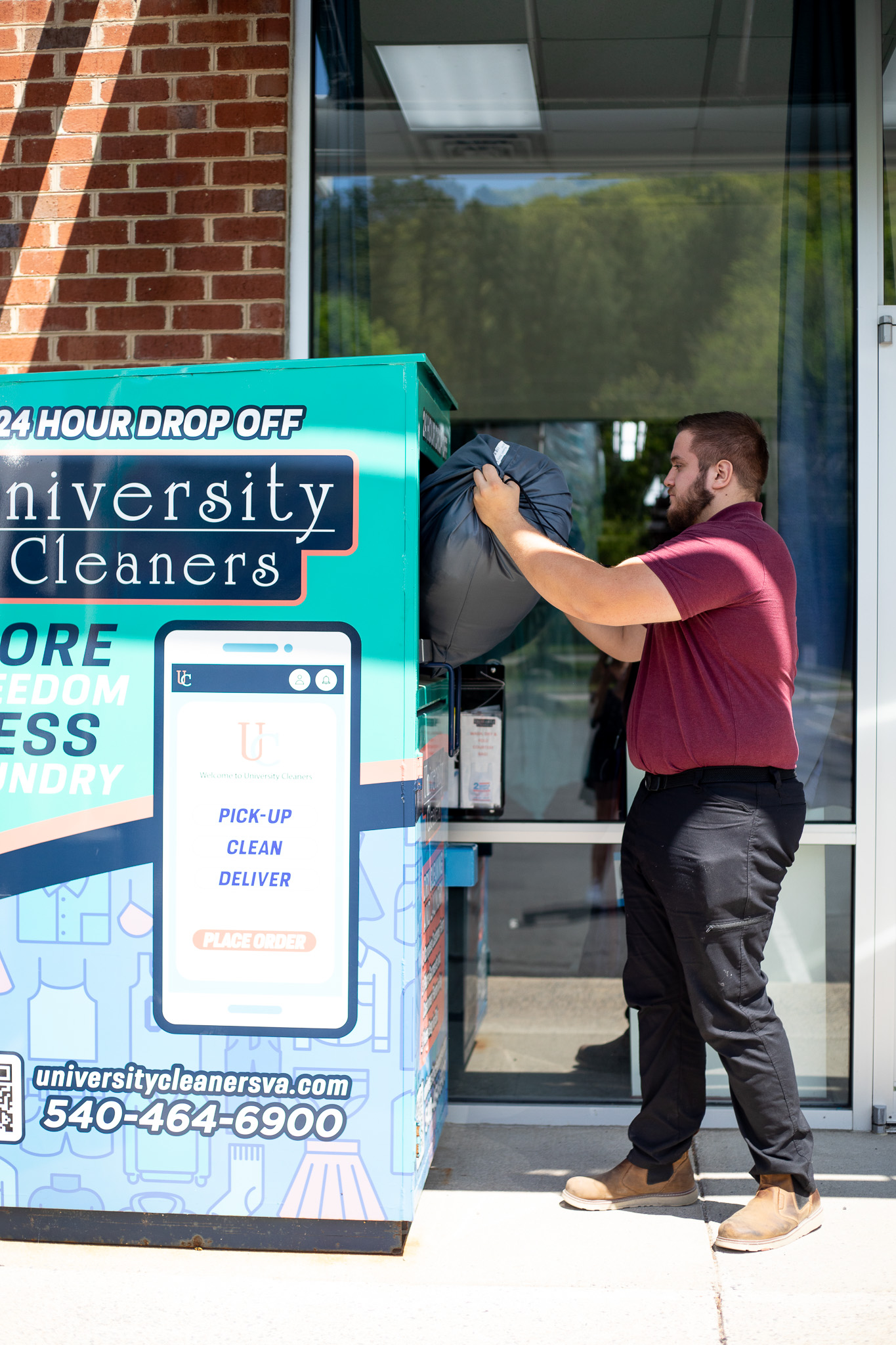 A man places a laundry bag into an outdoor University Cleaners 24-hour drop-off kiosk next to a storefront.