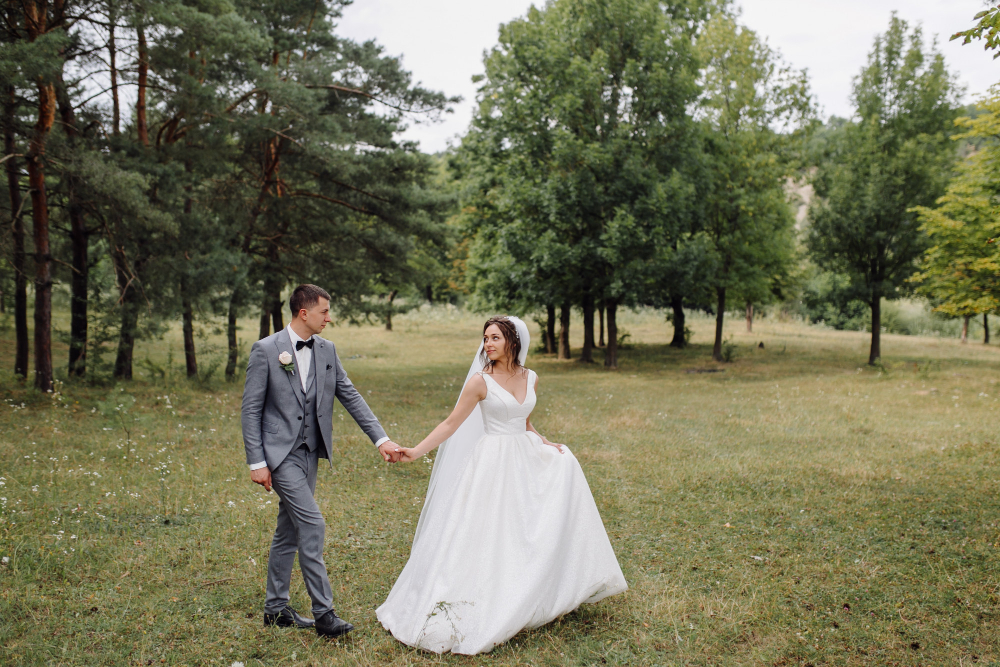 A bride in a white gown and a groom in a gray suit hold hands and walk together outdoors on a grassy area surrounded by trees.