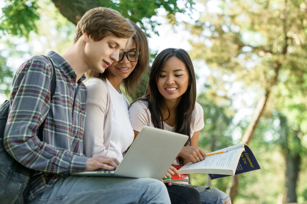 Three students sitting outdoors, looking at a laptop and a textbook together, surrounded by trees.