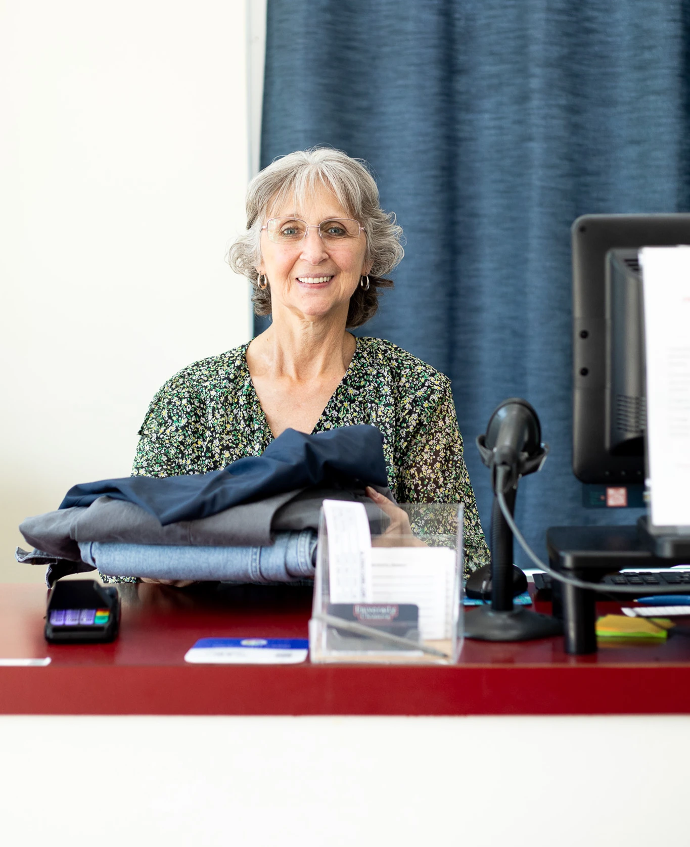 An older woman stands behind a counter with folded clothes, a computer, and a card reader, smiling at the camera.