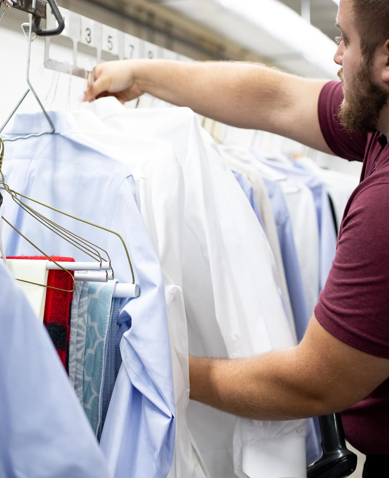 A person in a maroon shirt hangs freshly cleaned dress shirts on a rack at a dry cleaning facility.