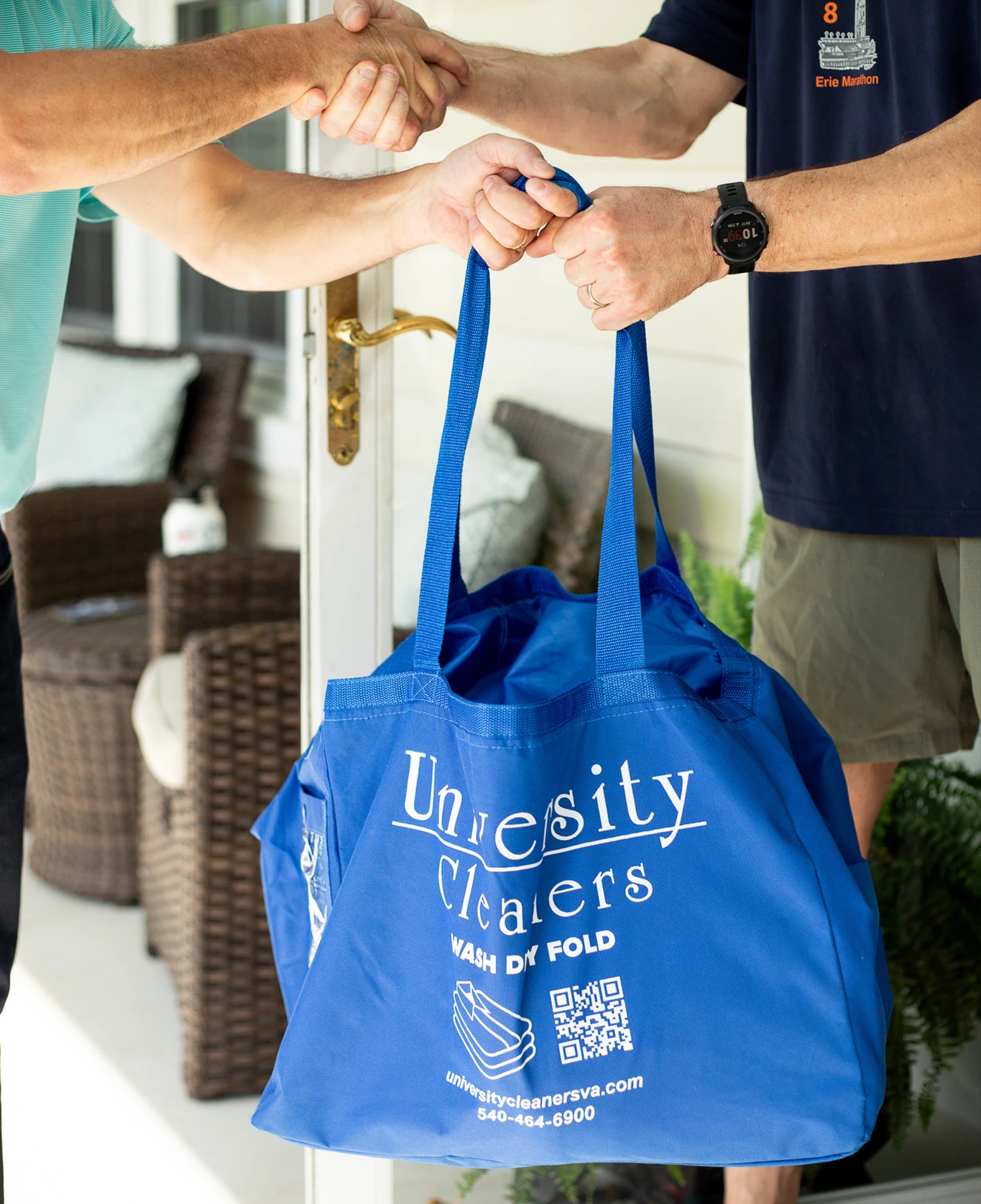 Two people shake hands as one hands over a blue "University Cleaners" laundry bag at a porch entrance.