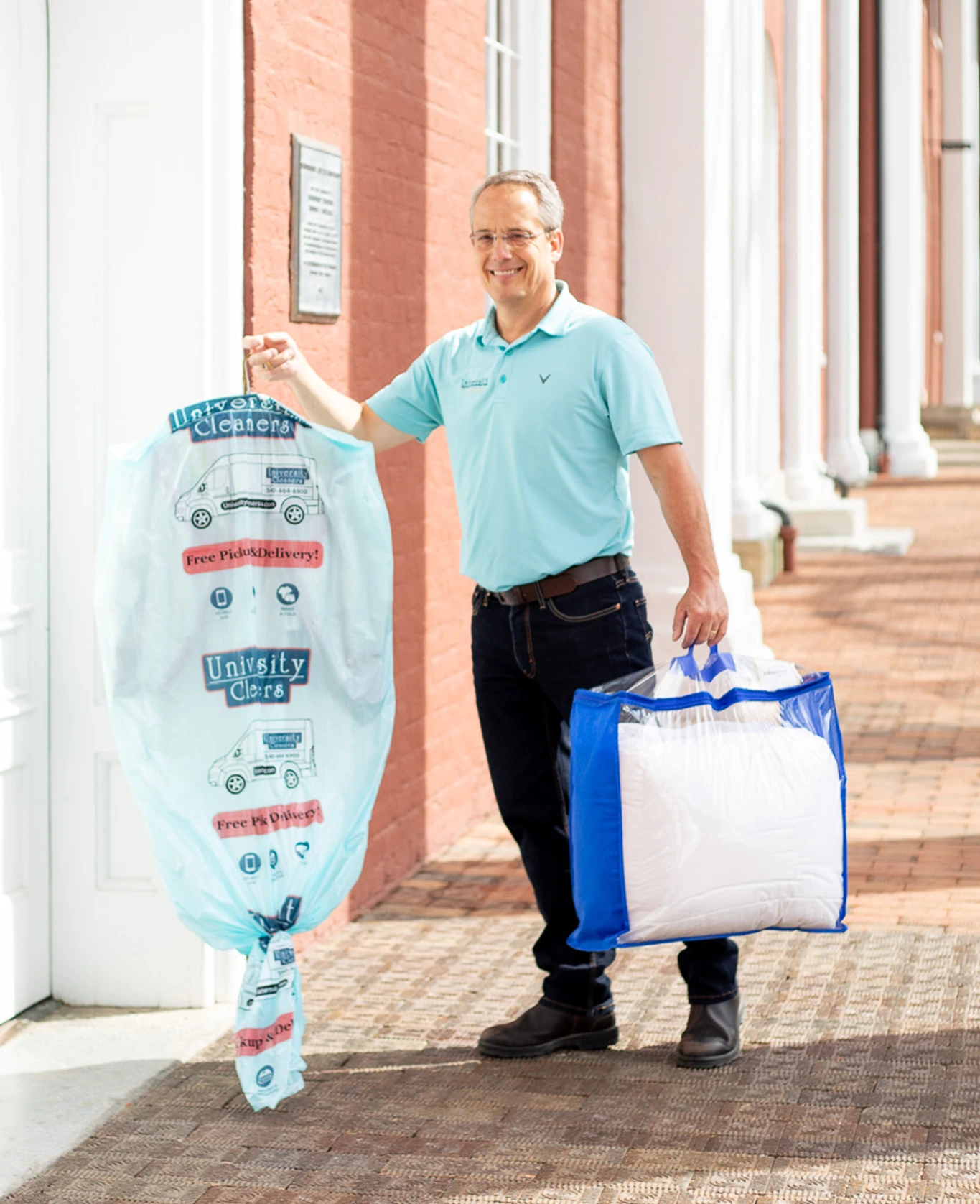 A man in a light blue polo shirt holds a garment in a plastic dry cleaning cover with one hand and a blue bag with a pillow in the other, standing outside a brick building.