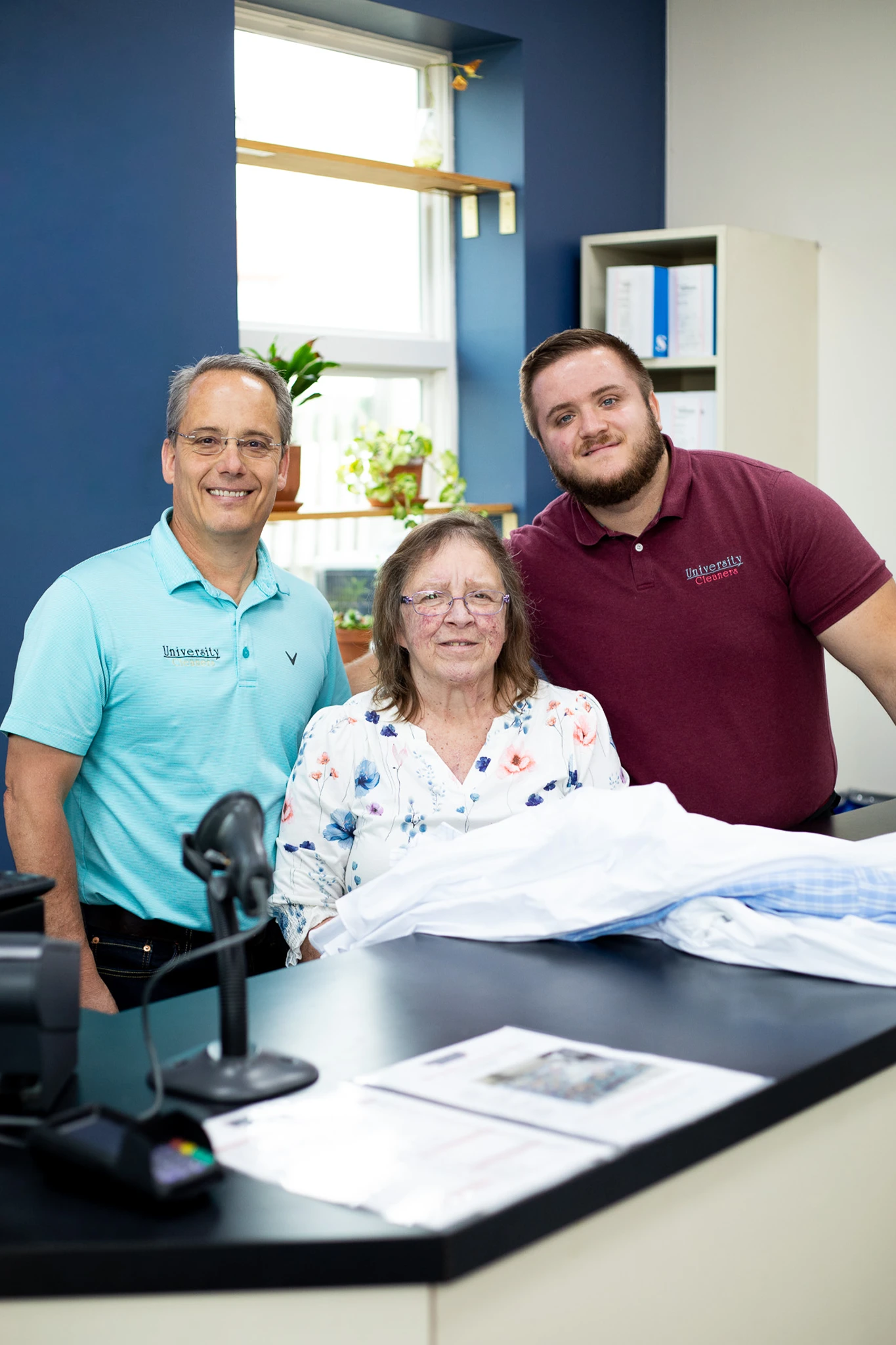 Three people, two men and an older woman, stand together behind a counter in an office environment, smiling at the camera.