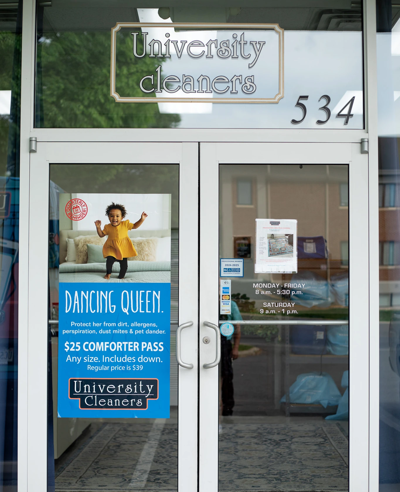 Glass entrance door of University Cleaners with street number 534 and business hours sign; a poster advertises a $25 comforter cleaning special with a child dancing.