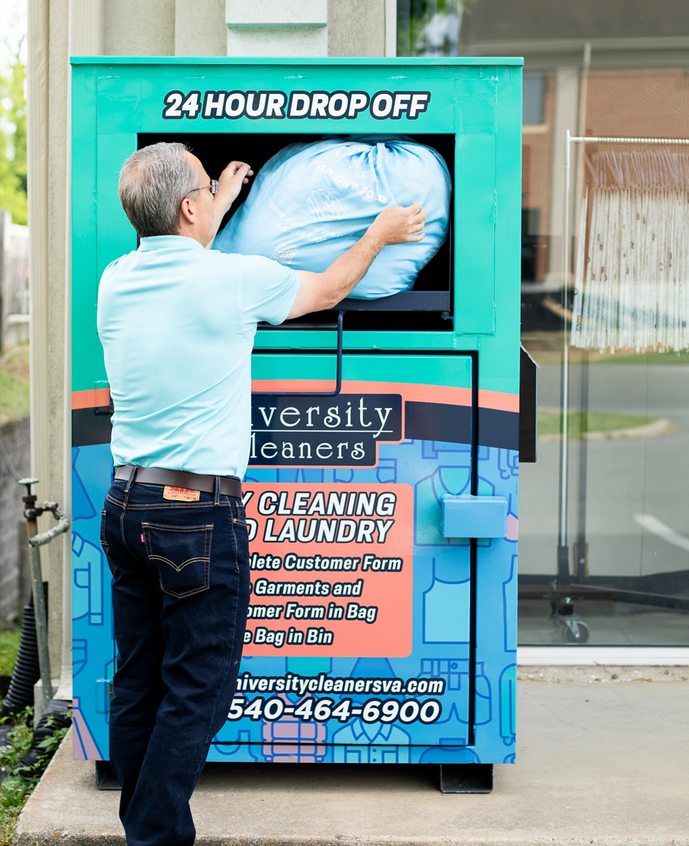 A man places a large blue laundry bag into a 24-hour drop-off bin outside a dry cleaning business.