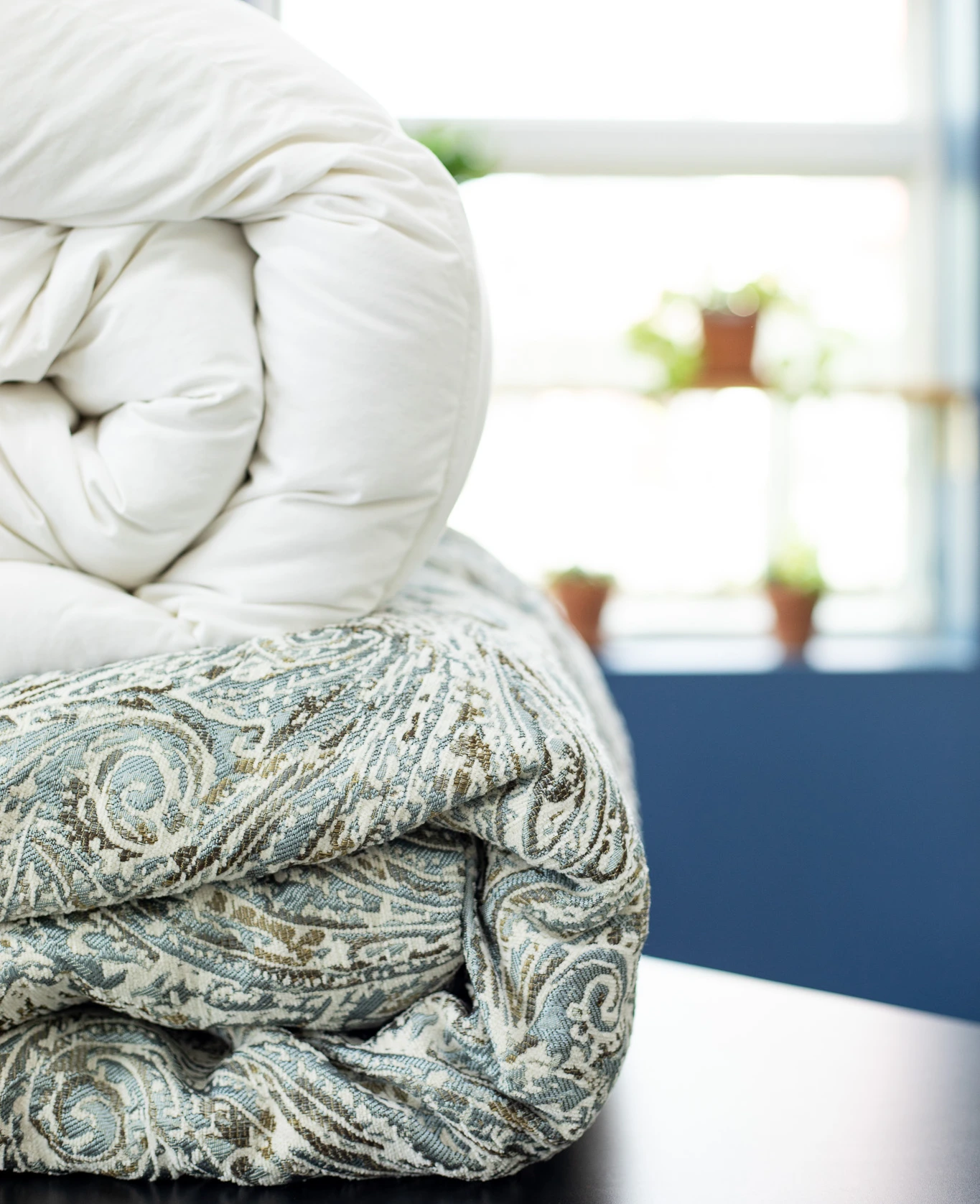 A close-up of a rolled white comforter stacked on top of a folded, patterned blue and beige blanket, with a blurred window and potted plants in the background.