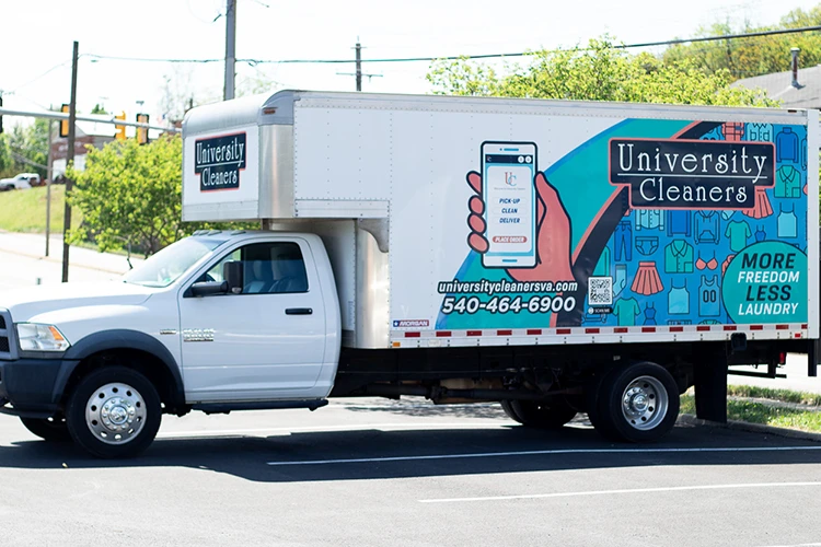 A University Cleaners box truck parked in a lot, featuring company branding, contact info, and laundry service graphics on the side.