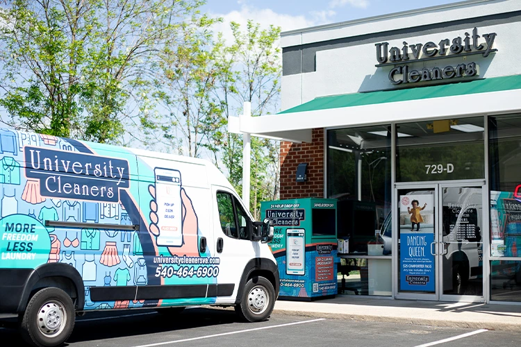 A University Cleaners van is parked outside a University Cleaners storefront, with branded signs, vending machines, and glass doors visible.
