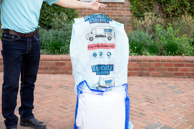 A person stands next to a large plastic laundry bag labeled "University Cleaners" on a brick pathway, with a brick wall and greenery in the background.
