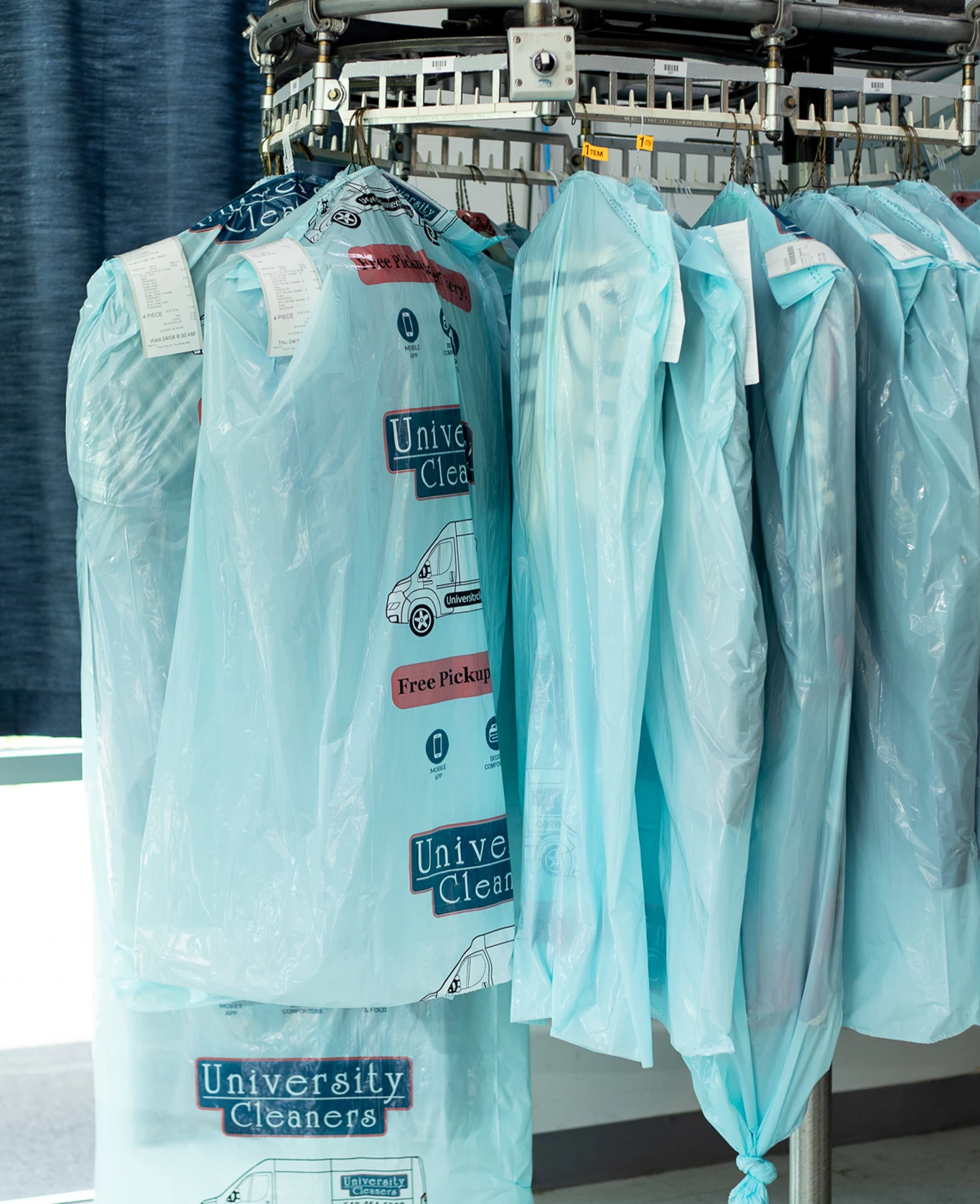Clothes covered in blue plastic bags hang on a rotating dry cleaning rack, with "University Cleaners" printed on the bags.